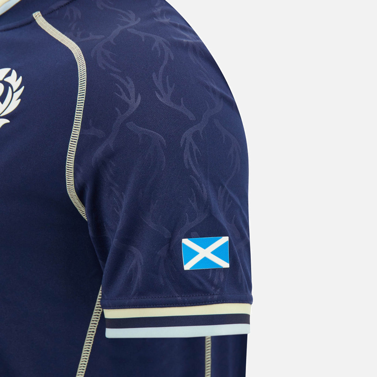 Sleeve detail from the Scotland SRU 26/27 Replica Home Jersey by Macron, featuring a navy sports jersey sleeve with a subtle antler pattern, cream trim, and a small blue and white Scottish flag patch on the lower sleeve—design elements inspired by the official Scotland rugby kit. This high-quality piece mirrors the craftsmanship of an authentic Macron Scotland jersey.