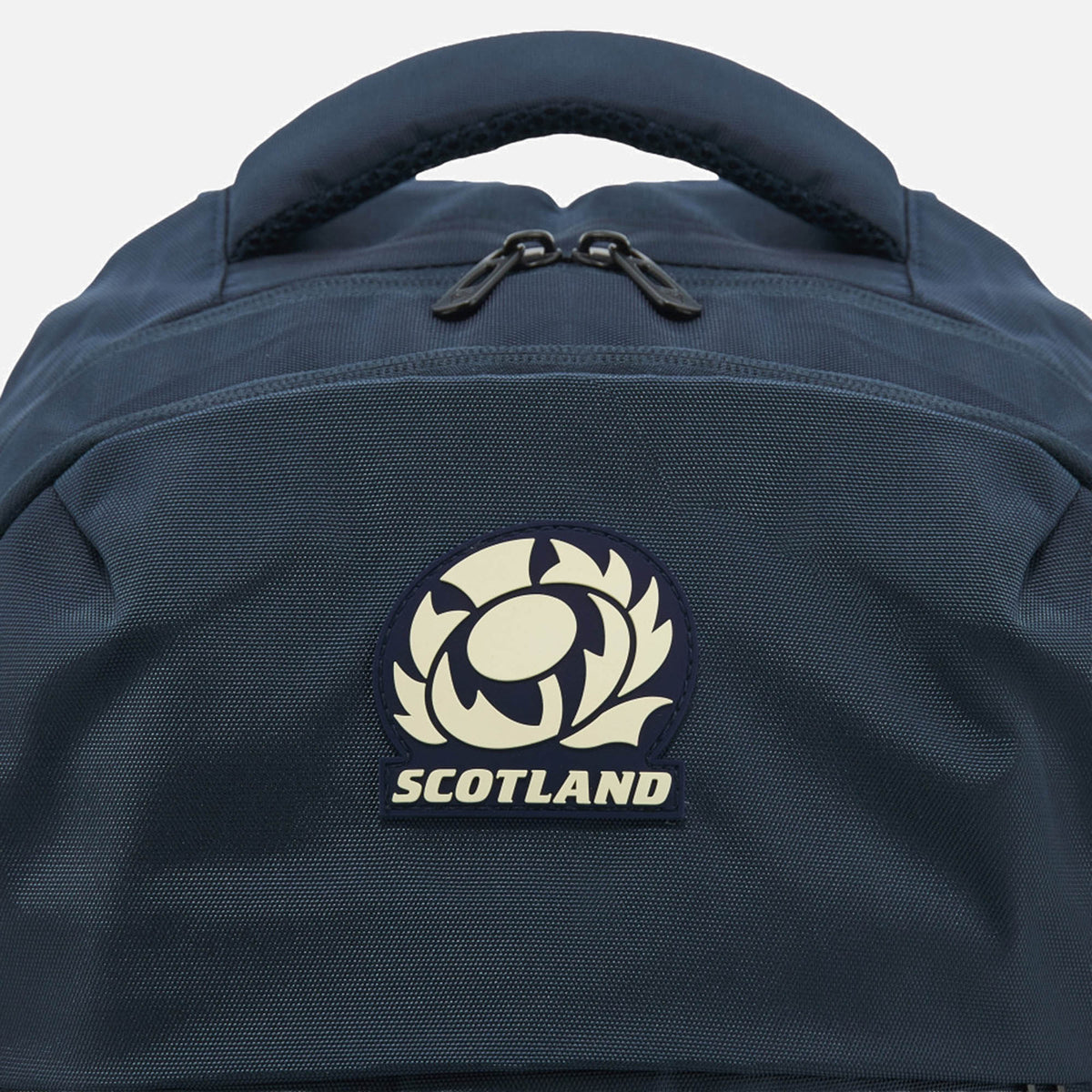 Close-up of the Scotland SRU 26/27 Backpack by Macron in blue, showcasing a prominent Scotland Rugby emblem patch centered on the front pocket. The backpack features a top carry handle and two visible zipper pulls—making it the perfect sports backpack for loyal Scotland rugby supporters.