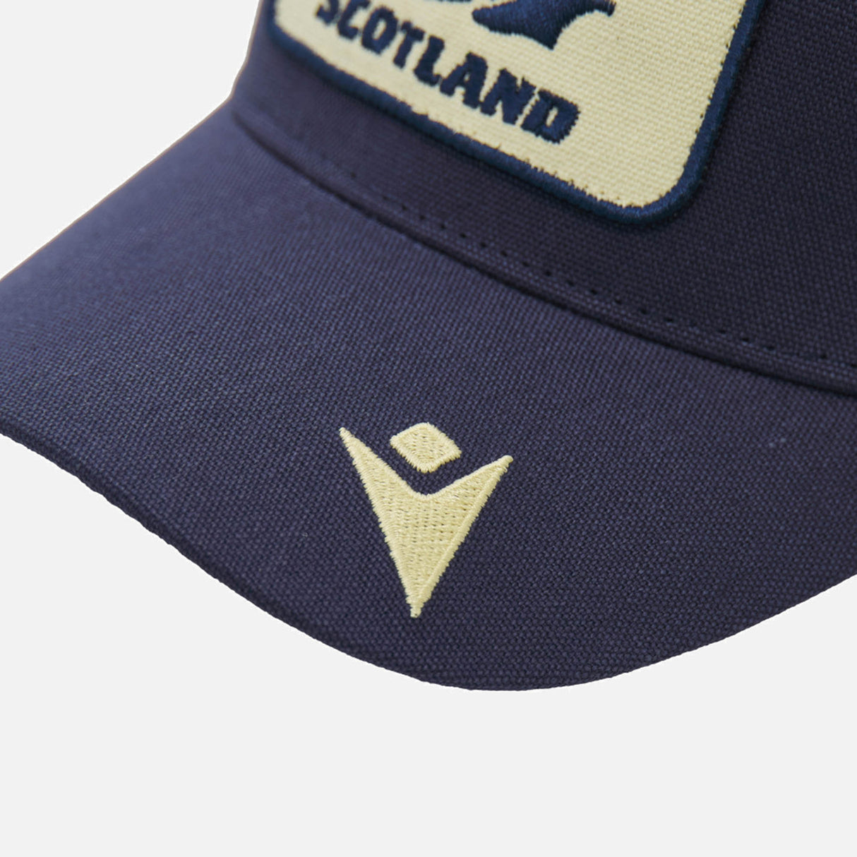 Close-up of the Scotland SRU 26/27 Trucker Cap by Macron in navy blue, featuring a yellow "Scotland" patch and a yellow embroidered logo on the bill.