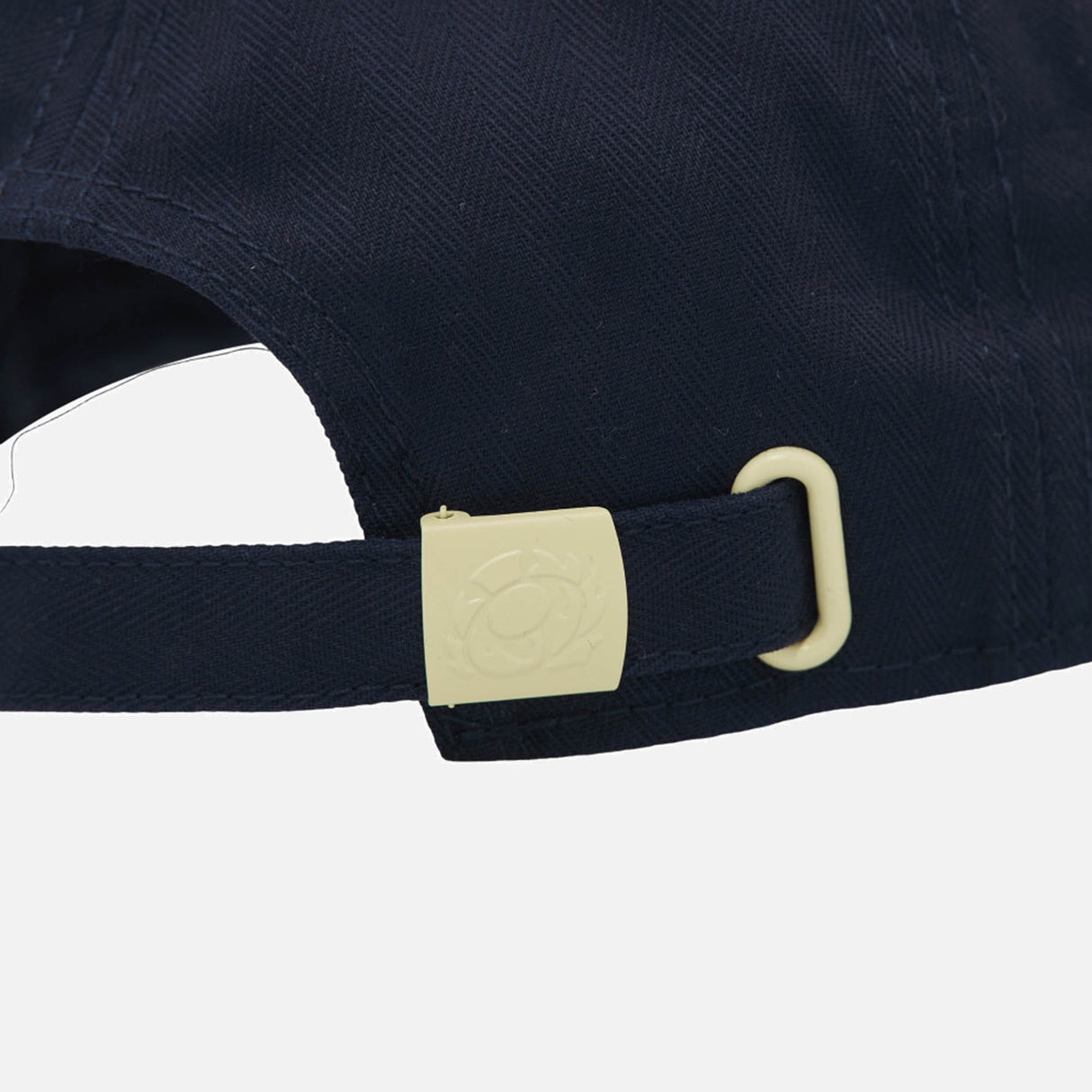 Close-up of the back of the Scotland SRU 26/27 Thistle Baseball Cap by Macron in navy blue, featuring an adjustable fabric strap and a cream-colored metal clasp with an embossed circular design.