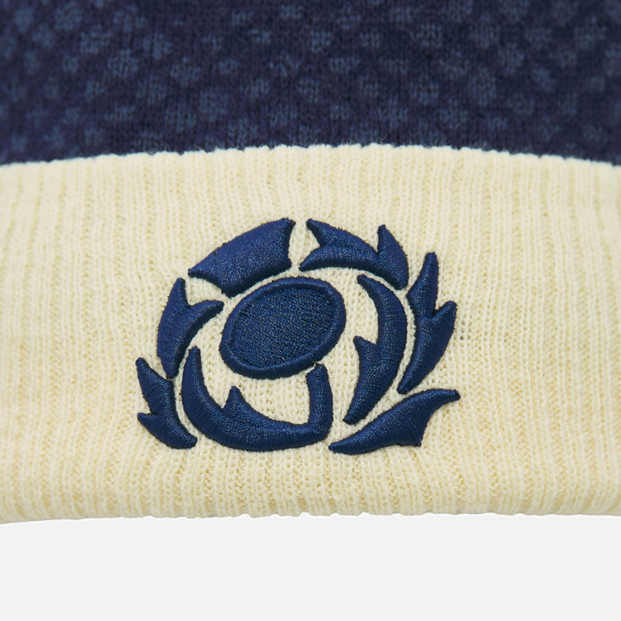 The Scotland SRU 26/27 Pom-Pom Beanie by Macron features a navy and cream knit design with an embroidered thistle emblem on the cream section, symbolizing Scottish rugby.