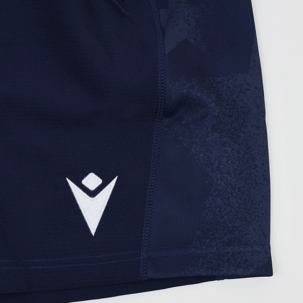 Close-up of the lower part of USA Rugby 24/25 Home Shorts by Macron in navy blue, featuring a white embroidered geometric Macron logo near the hem.