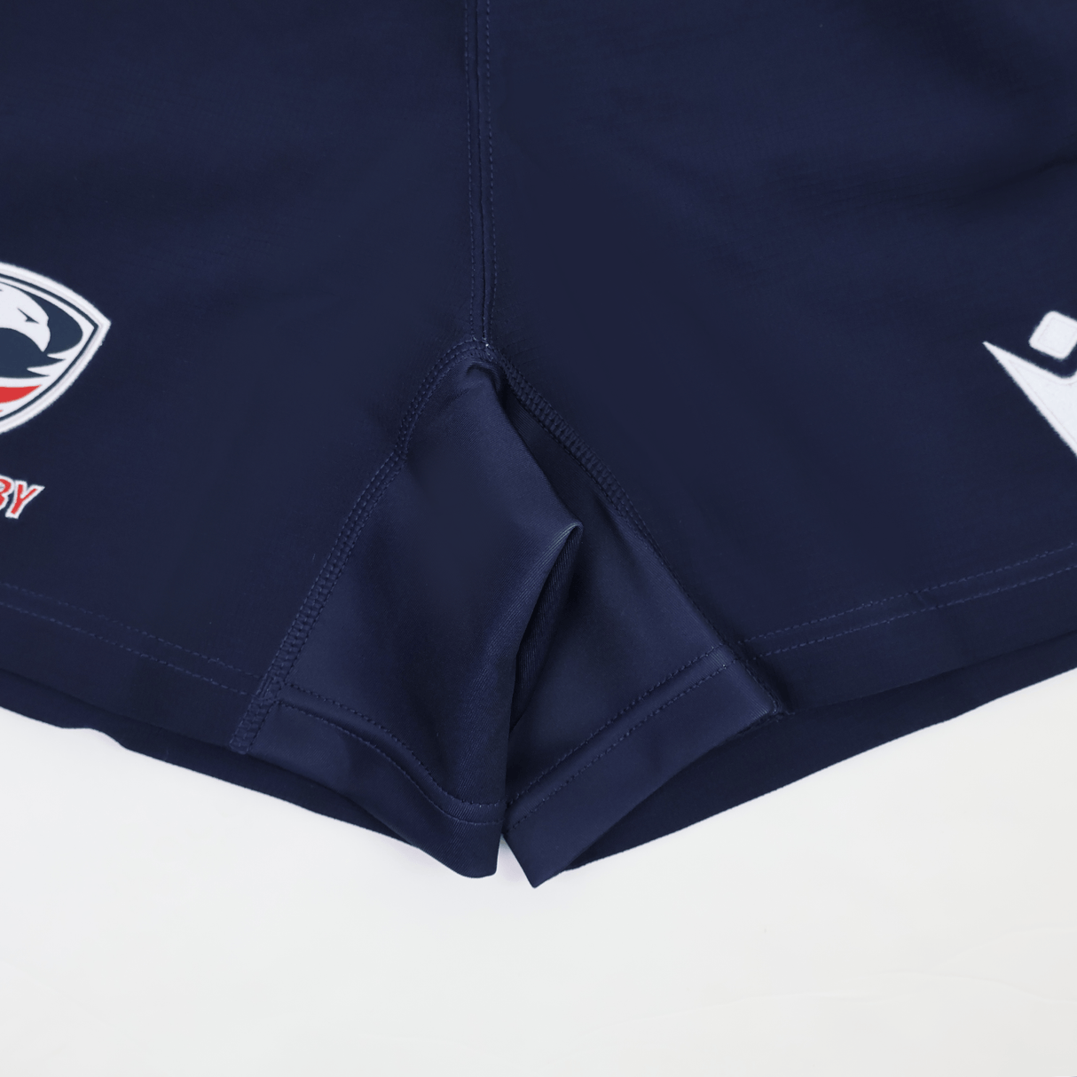 Close-up of USA Rugby 24/25 Home Shorts by Macron in navy blue, featuring visible stitching and two logo patches near the hem, designed for moisture-wicking performance.