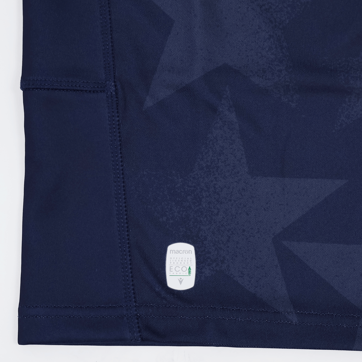 Close-up of navy blue fabric with subtle star patterns, visible stitching, and a small white "macron ECO" label near the hem—part of the Youth USA Rugby 24/25 Home Jersey by Macron.