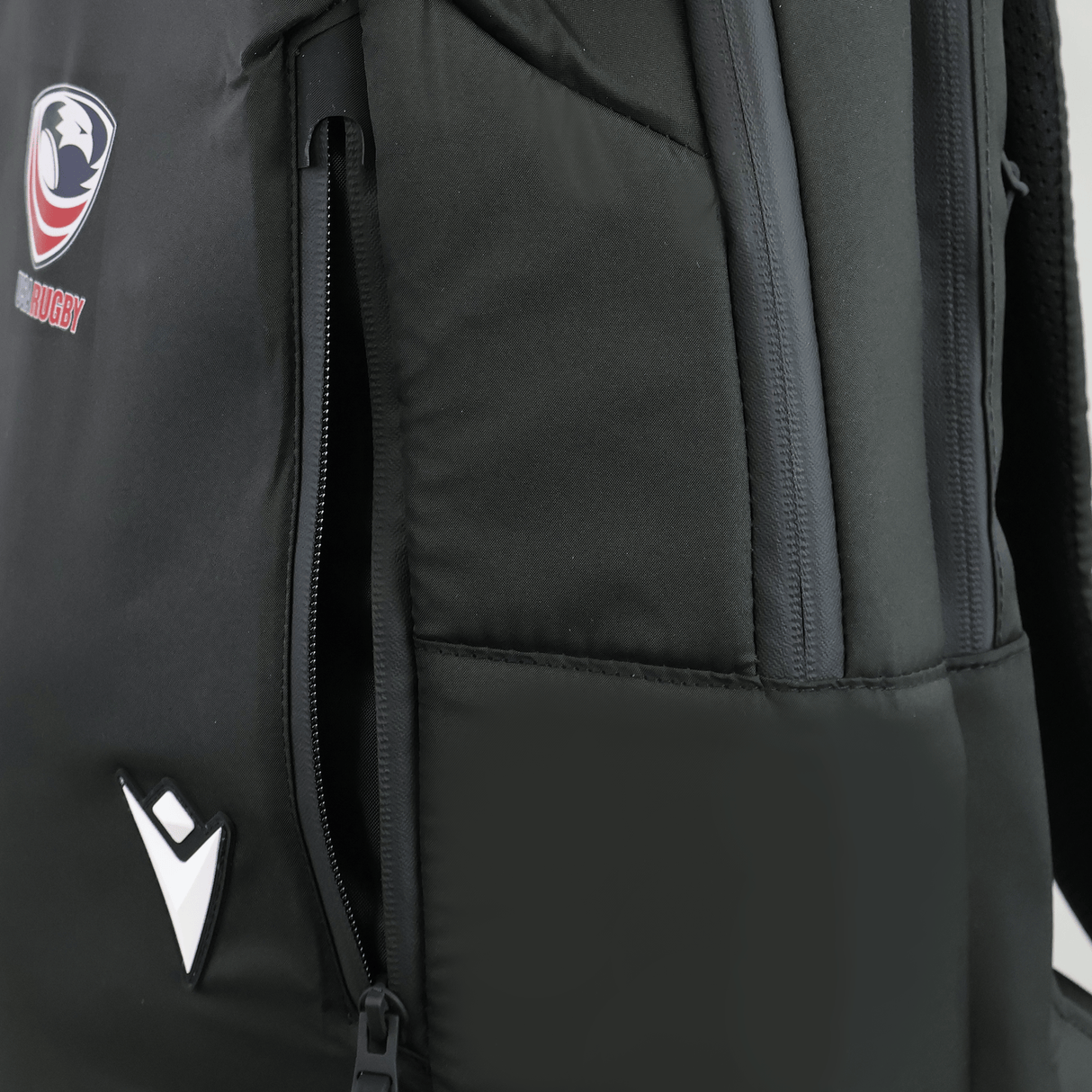 Close-up of the USA Rugby 25/26 Staff Backpack by Macron in black, showing a partially unzipped side pocket, rugby logo, and a white geometric emblem on the front.