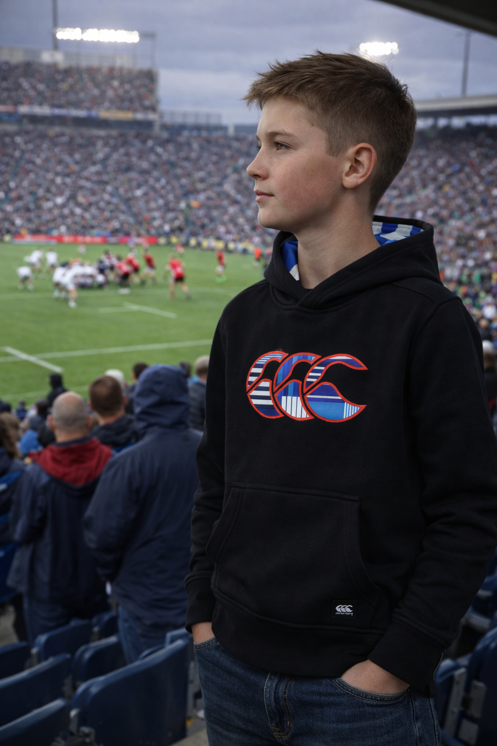 A boy wearing the Canterbury Youth Uglies Logo Hoodie stands in a stadium, watching a rugby game unfold before a lively crowd, fully immersed in the vibrant atmosphere of rugby style.