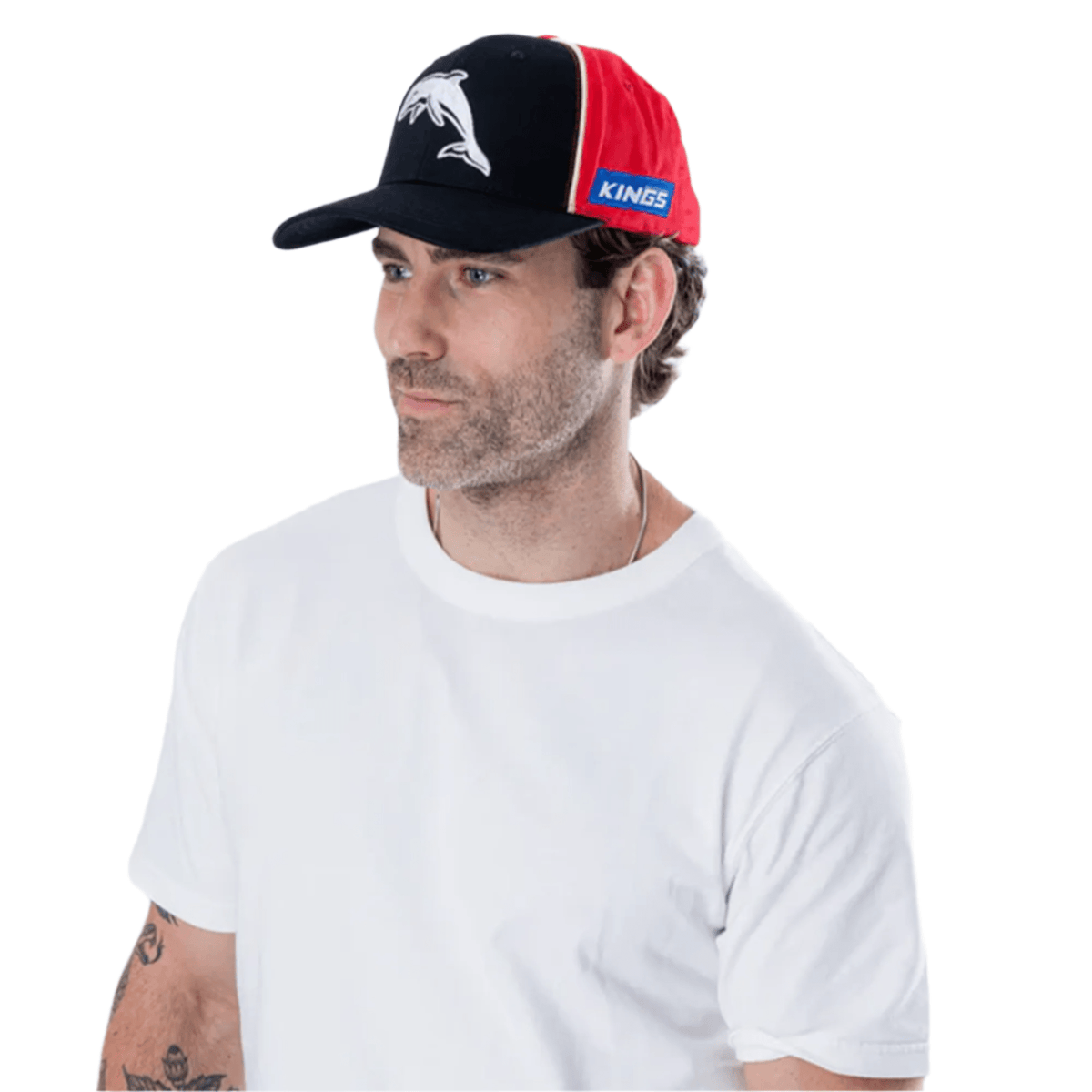 A man in a white t-shirt and a Dolphins NRL 25/26 Media Cap by Classic Sportswear, adorned with black, red, and blue colors and the NRL logo, gazes thoughtfully to the side.