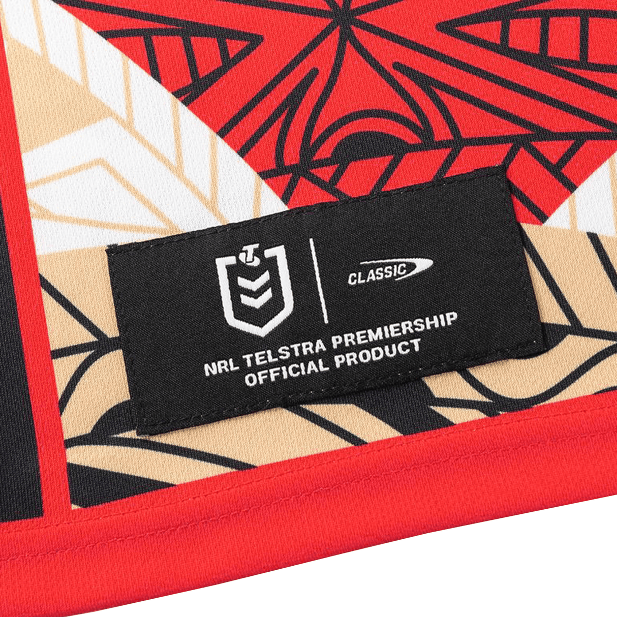 A fabric label reading "NRL Telstra Premiership Official Product" is attached to the Dolphins Indigenous NRL 25/26 Replica Jersey by Classic Sportswear, featuring a geometric red, black, beige, and white pattern.