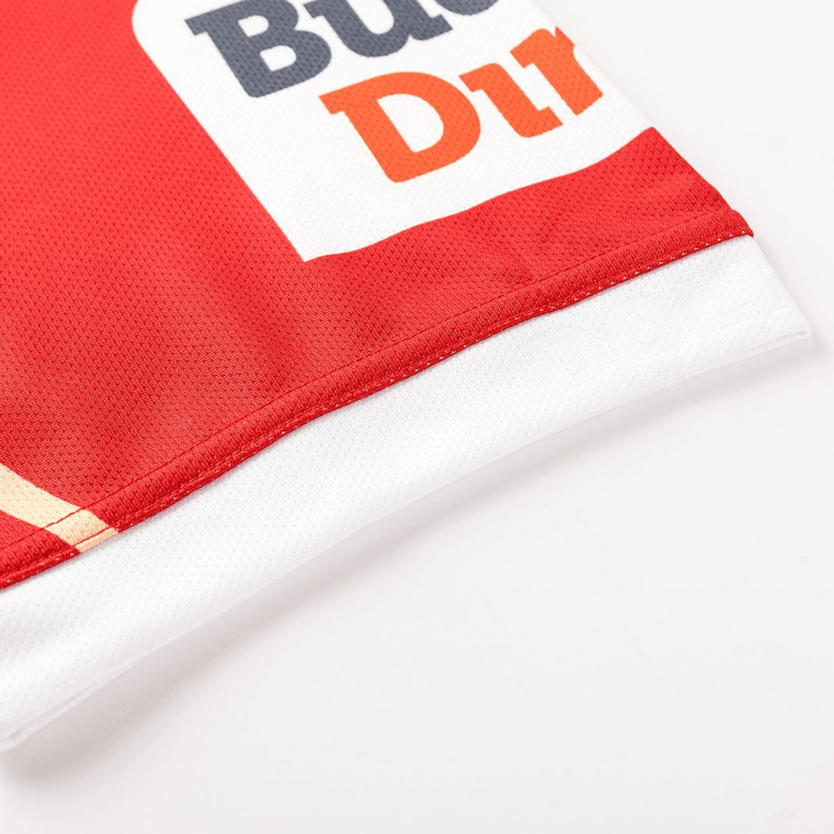 Close-up of the edge of a red and white Dolphins NRL 26/27 Replica Home Jersey by Classic Sportswear, showing partial printed text and stitched seams on a white background.