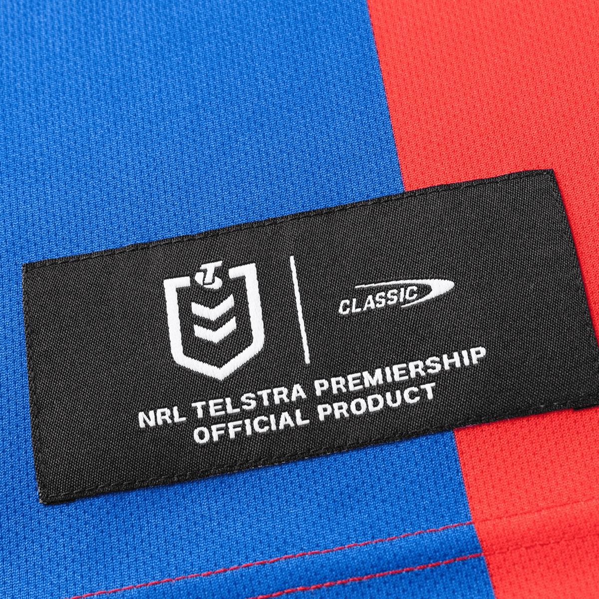 A black fabric label with white text reading "NRL Telstra Premiership Official Product" appears on the blue and red Knights NRL 26/27 Replica Home Jersey by Classic Sportswear.
