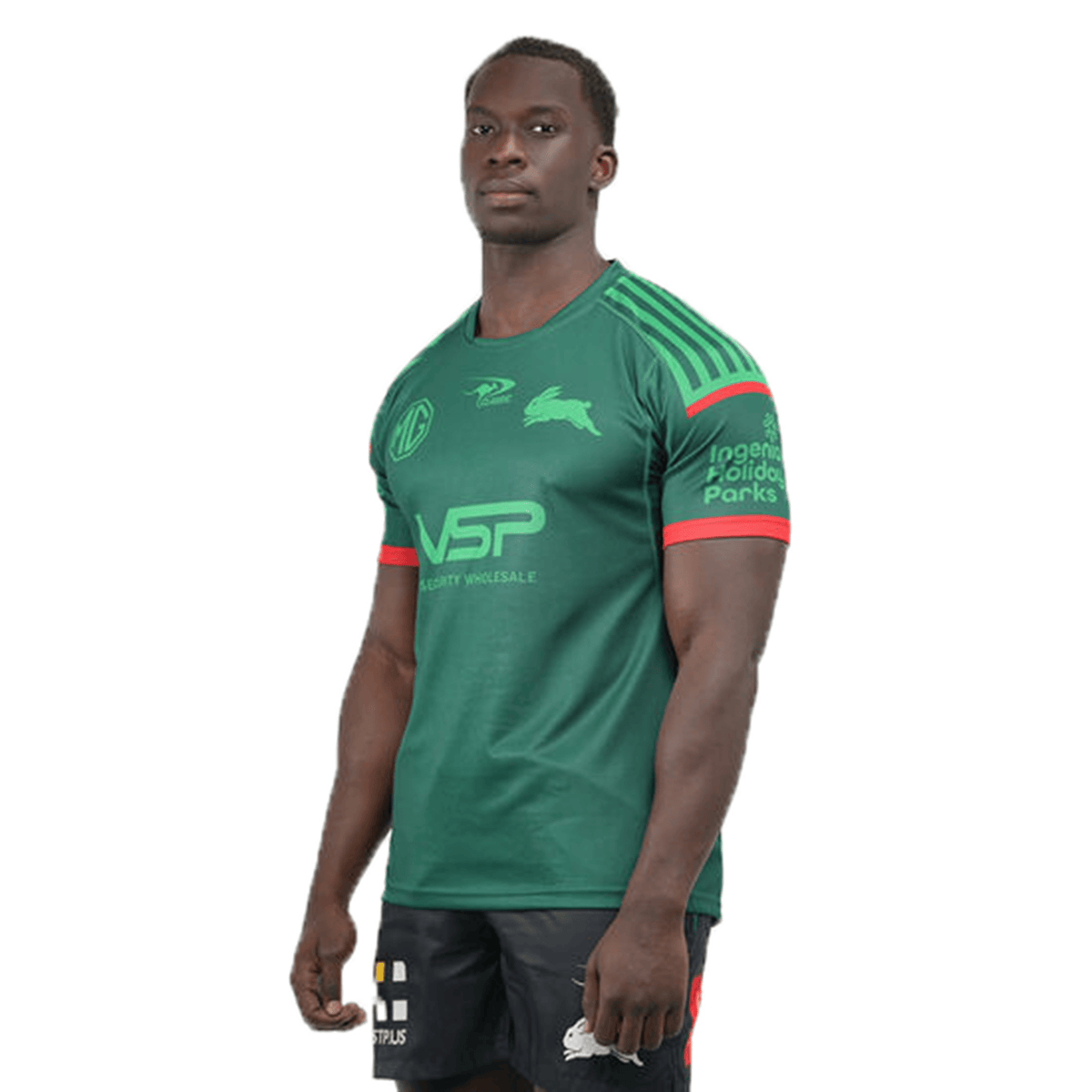 A man in a Classic Sportswear Rabbitohs NRL 25/26 Special Edition Captain's Run Jersey with logos and black shorts stands against a white background.