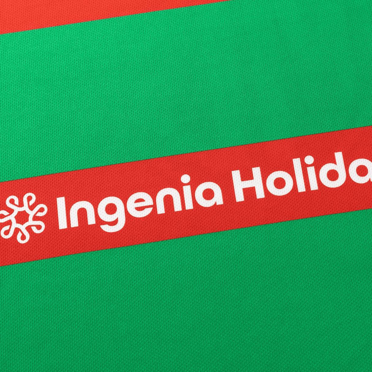 Close-up of a red and green fabric with "Ingenia Holiday" and a star logo on the red stripe, from the Rabbitohs NRL 26/27 Replica Home Jersey by Classic Sportswear.