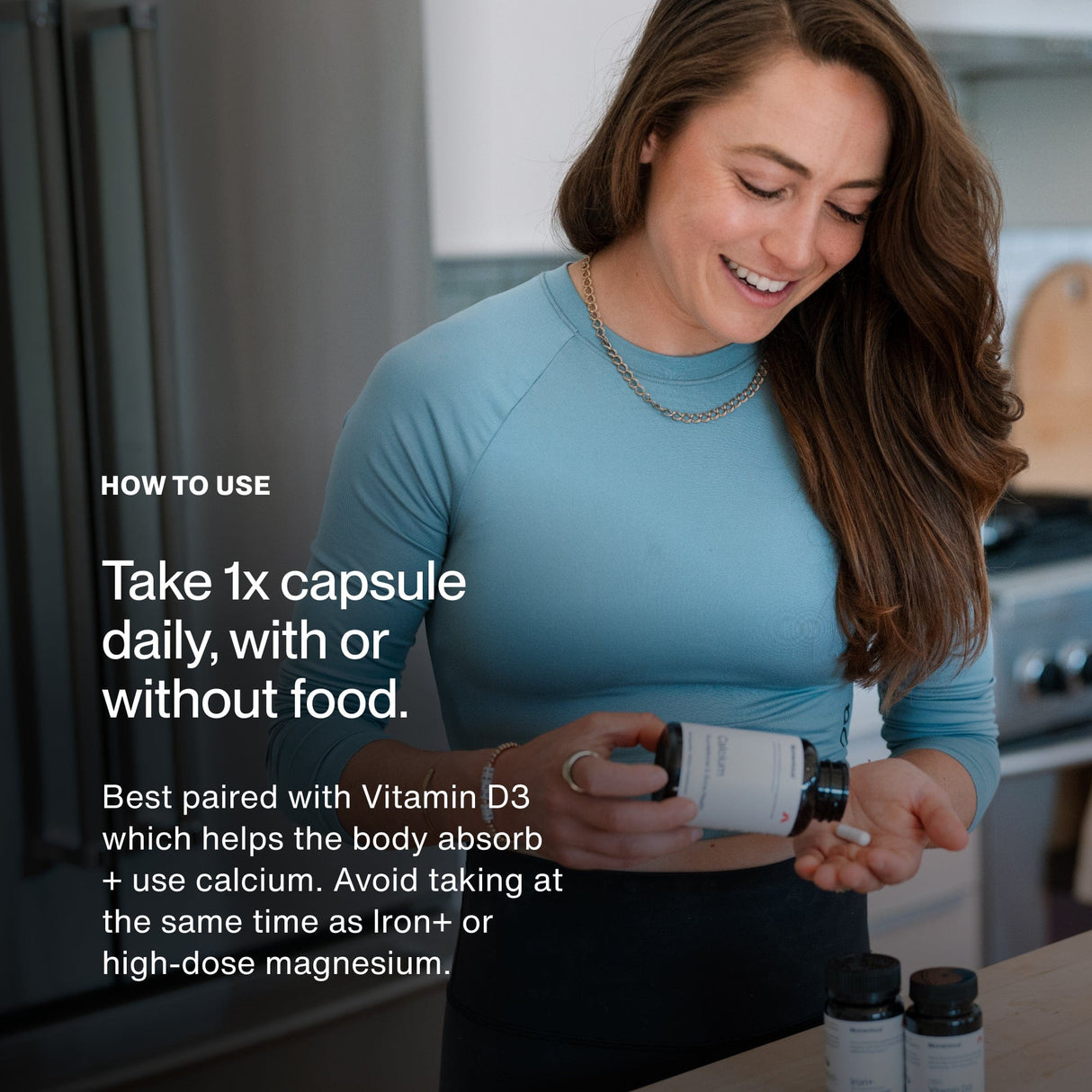 A woman in a blue top stands in a kitchen, holding a bottle of Momentous Calcium by Momentous and pouring a capsule into her hand while usage instructions are printed nearby.