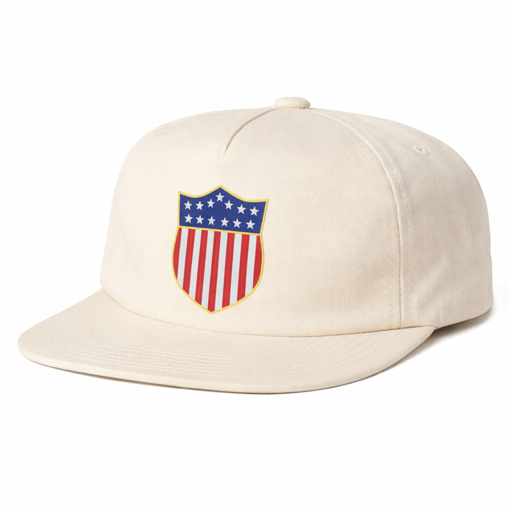 The EMB Weld USA Rugby Original Olympic Brushed Cotton Cap is beige and features a front shield emblem with blue and white stars above red and white vertical stripes.