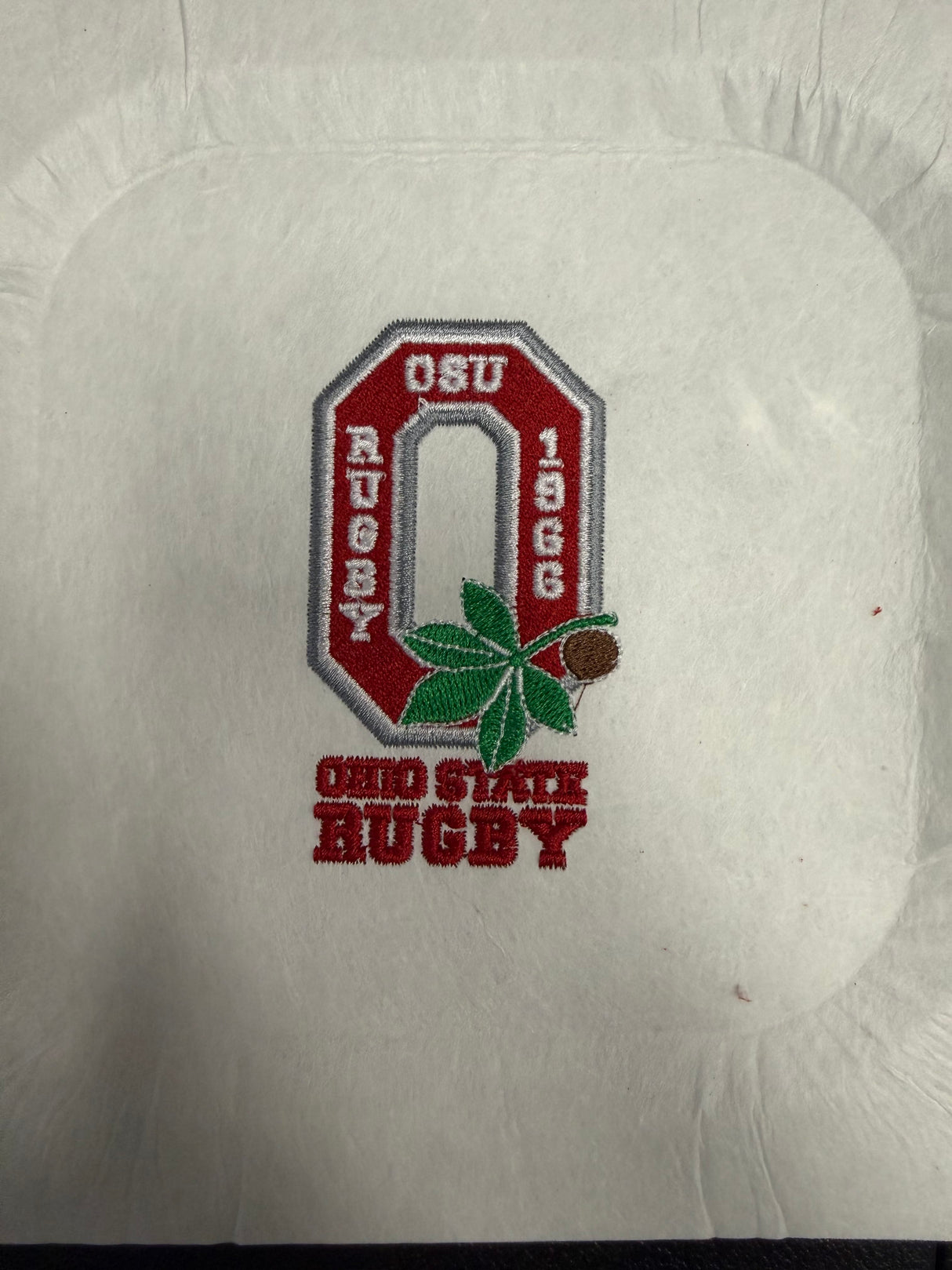 The Ohio State University Alumni EMB Logo by EMB World Rugby Shop features an embroidered red "O," buckeye leaf and nut, and the text "OSU Rugby 1966" and "Ohio State Rugby" on white fabric.