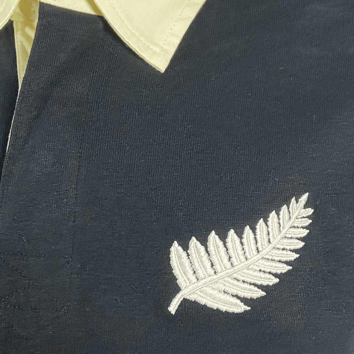 Close-up of the All Blacks 1983 Classic Rugby Shirt by Ellis Rugby, showcasing its dark fabric, yellow collar, and white embroidered fern design.