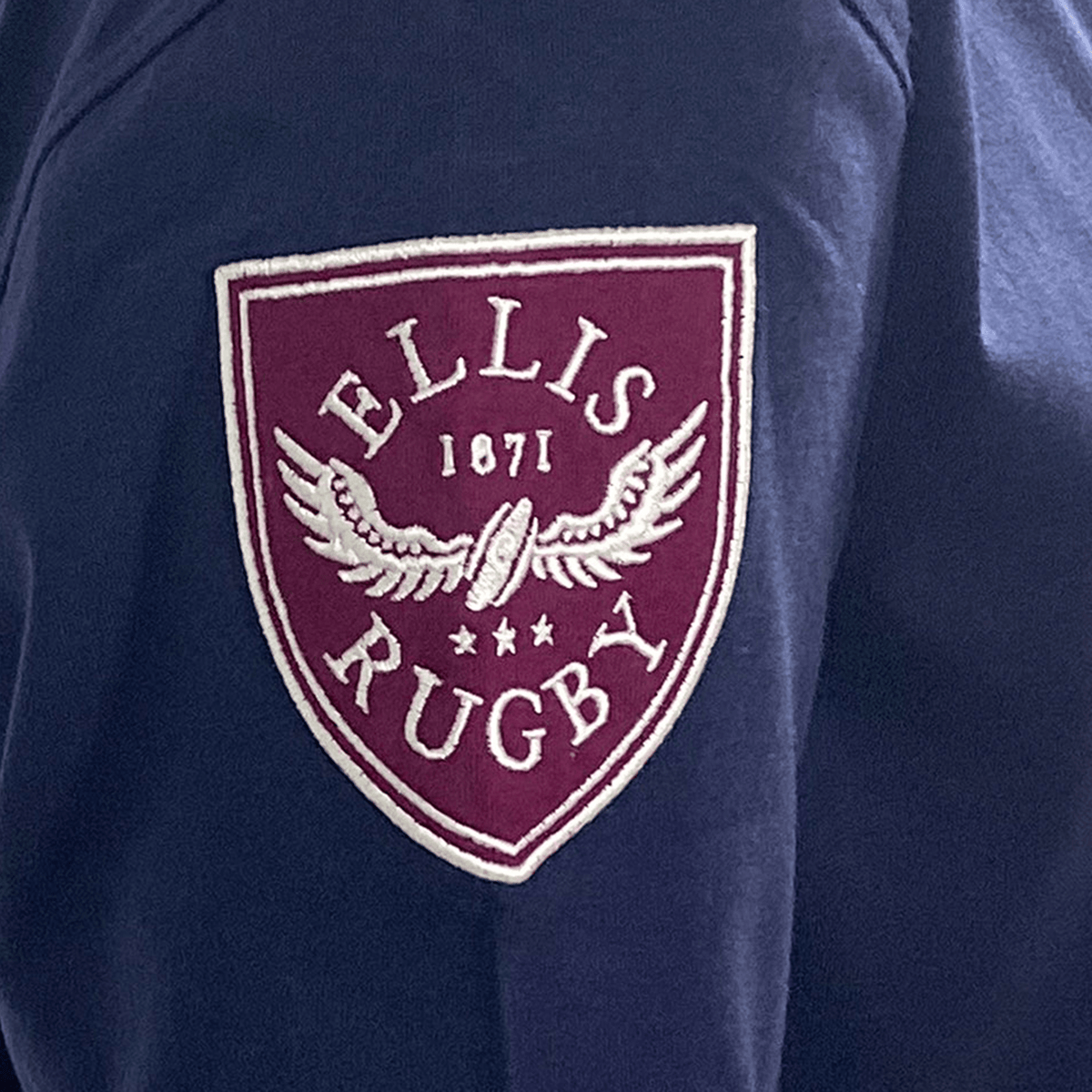 The Ellis Rugby England Rugby 1924 Grand Slam Shirt displays a maroon shield emblem on dark blue fabric, featuring "ELLIS 1871 RUGBY" and a winged rugby ball, symbolizing the Grand Slam victory spirit.