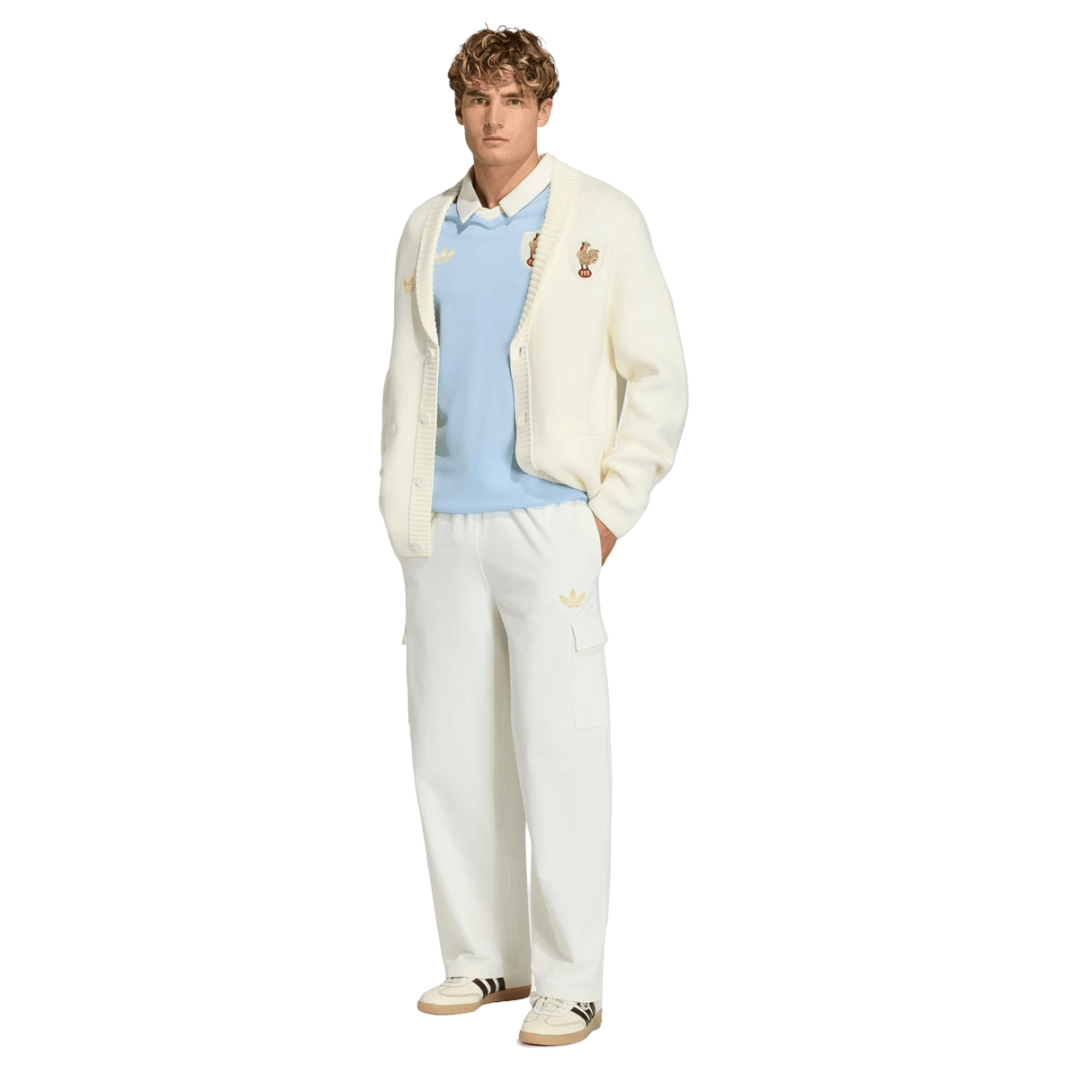 A man wears the adidas France FFR 120 Year Cardigan with embroidered patches, a light blue sweater over a collared shirt, white cargo pants, and cream sneakers with brown stripes, showcasing French rugby heritage.
