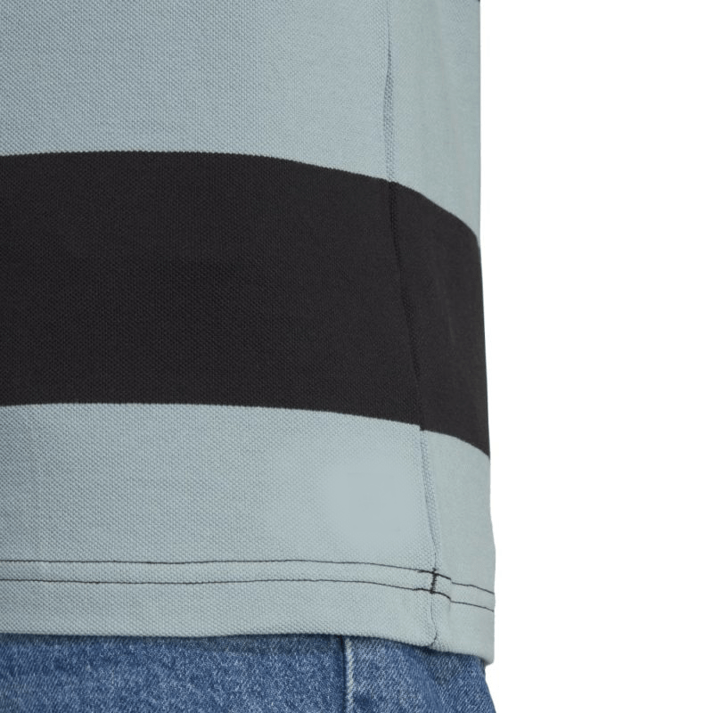 Close-up of the All Blacks Supporters Polo by adidas in light grey with a horizontal black stripe above the hem, reminiscent of rugby union hoops polo style, paired with blue denim jeans.