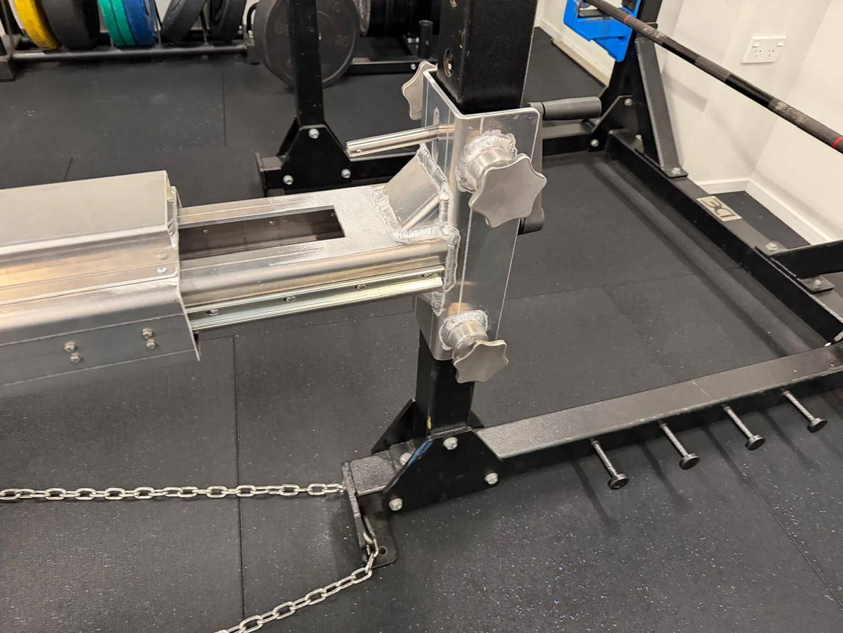 Close-up of the IronBack Iron Back Force Trainer gym machine attachment, featuring metal rods, adjustment knobs, and chains on a black rubber floor near a squat rack. Designed to help build isometric strength and enhance horizontal pushing power.