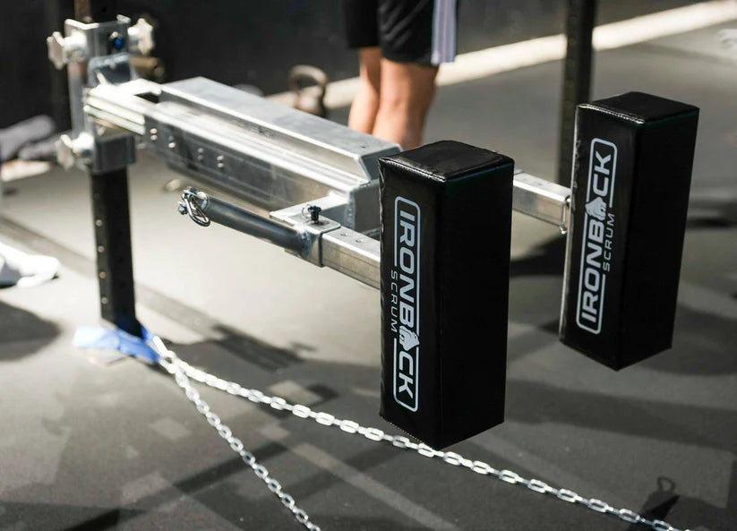 Close-up of the IronBack Force Trainer gym equipment with padded supports, branded "IronBack"; ideal for building horizontal pushing strength, with a person's legs and gym flooring visible in the background.