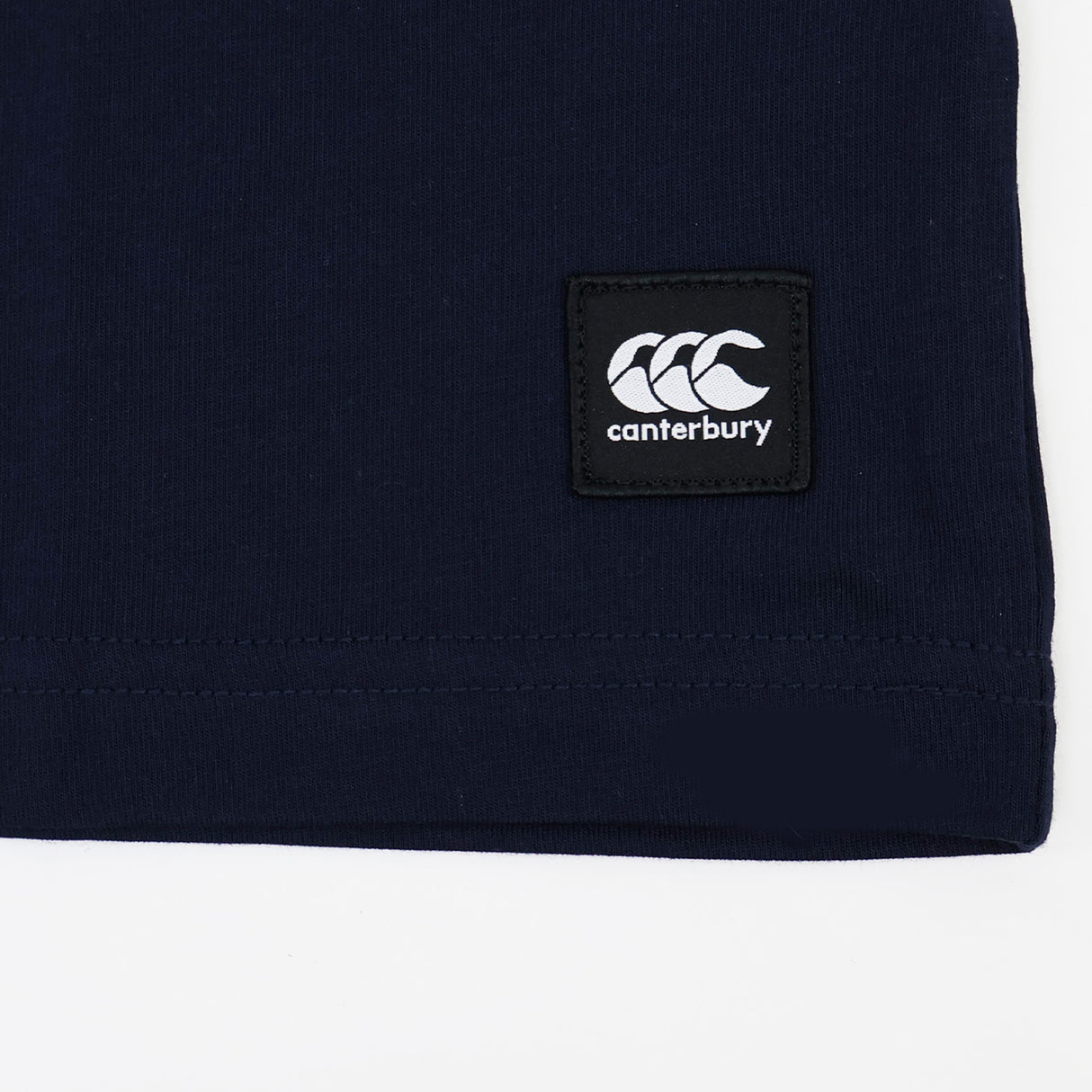 Close-up of a dark blue Canterbury Uglies Logo T-Shirt featuring a black and white Canterbury logo patch sewn near the hem, evoking the classic style associated with Canterbury rugby shirts.