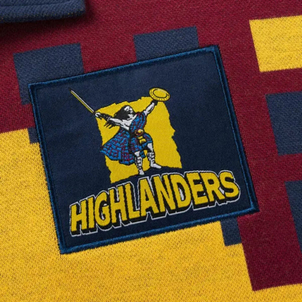 An embroidered patch displaying a kilted figure with a sword and shield, labeled "Highlanders," adorns the multi-colored fabric background of the Limited Edition Highlanders Super Rugby 25/26 Retro Jersey by Classic Sportswear.