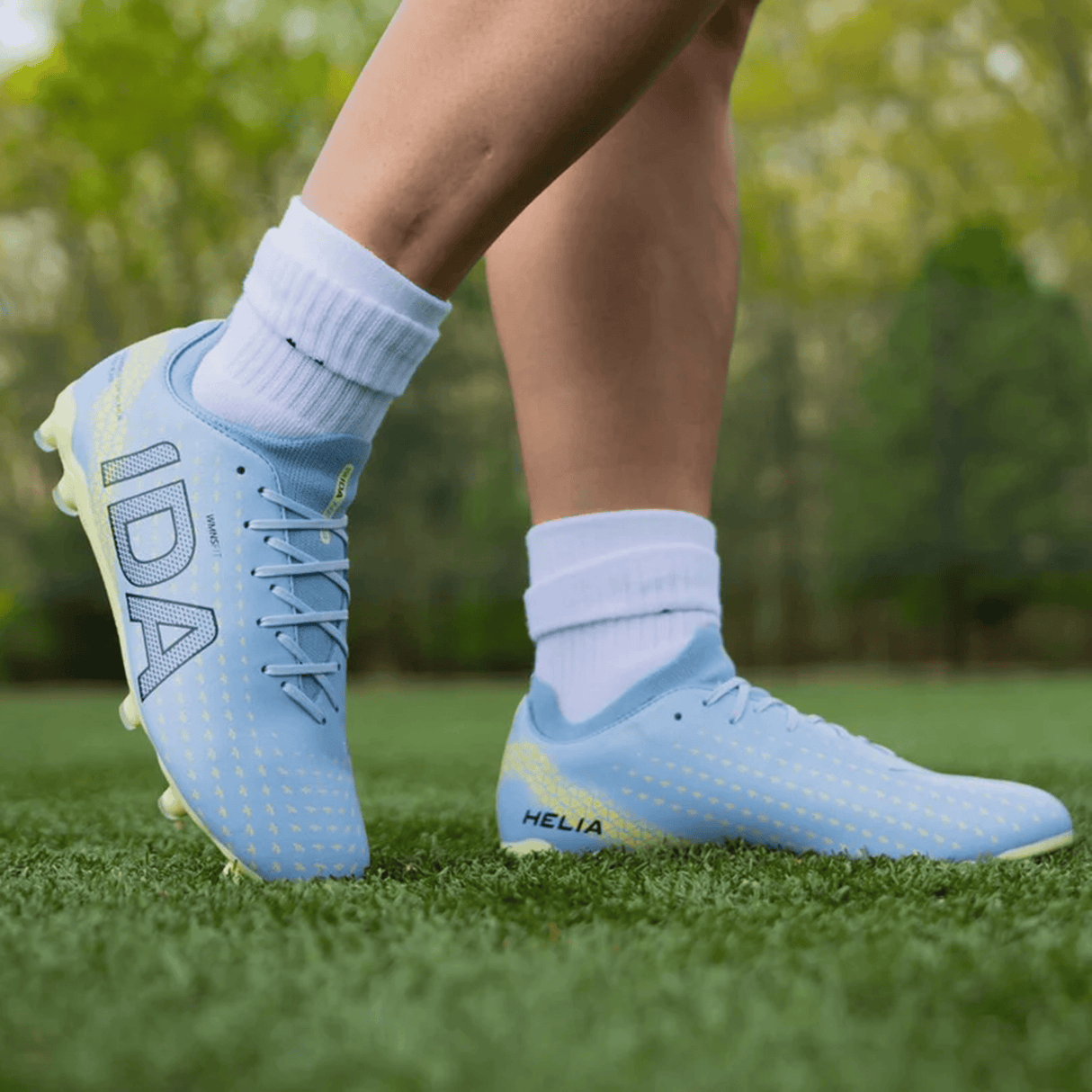 A person wearing IDA Sports’ IDA Helia Club FG – Light Blue women’s soccer cleats with white socks stands on green artificial grass, with blurred trees in the background.