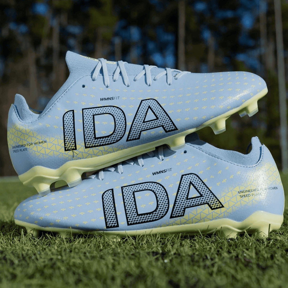 The IDA Sports IDA Helia Club FG - Light Blue soccer cleats for women feature yellow accents and bold "IDA" branding, stacked on a grassy field—ideal firm ground boots designed specifically for female players.
