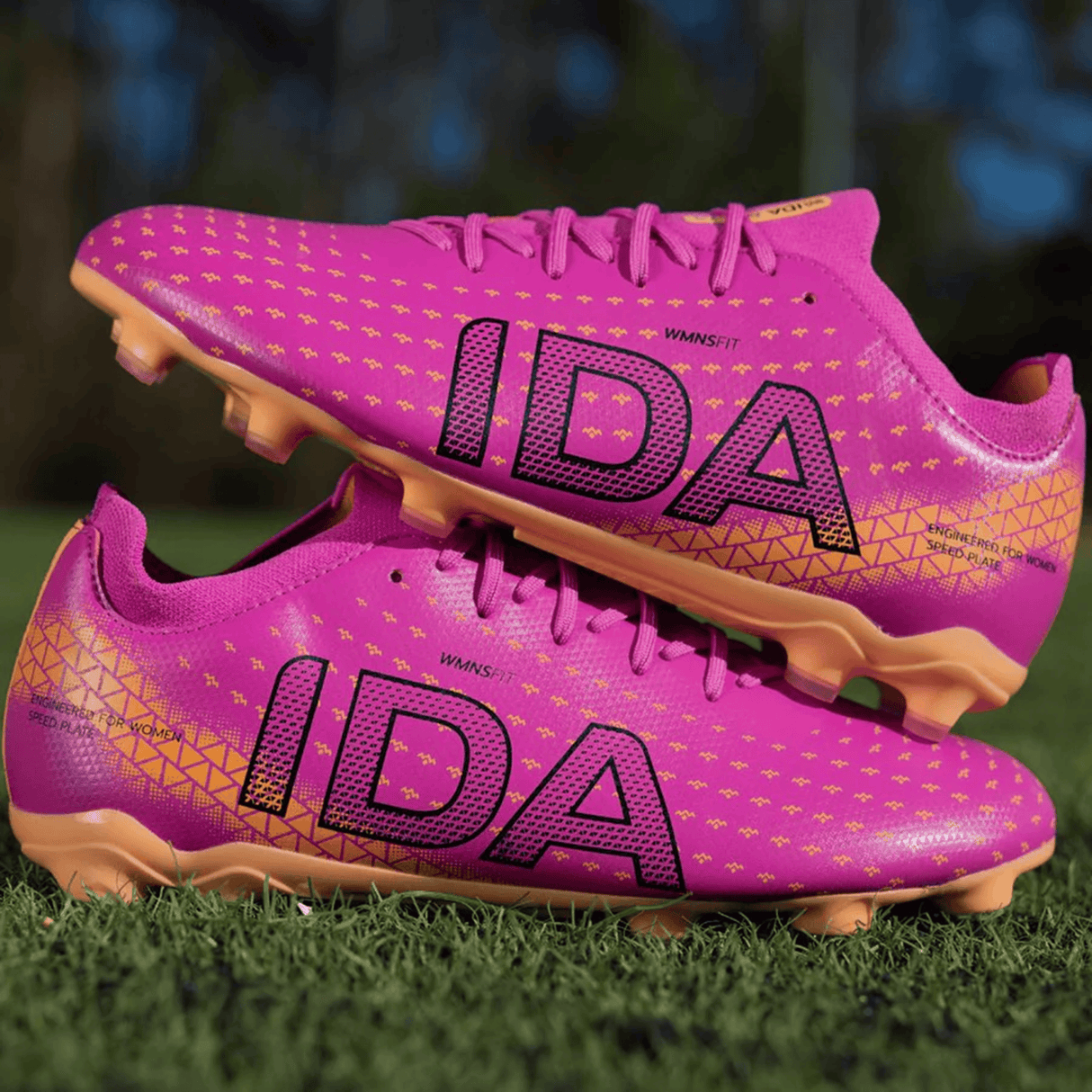 A pair of IDA Sports Helia Club FG women's soccer cleats in dark pink with gold accents and "IDA" printed in black, resting on green grass—these firm ground boots combine style and performance.