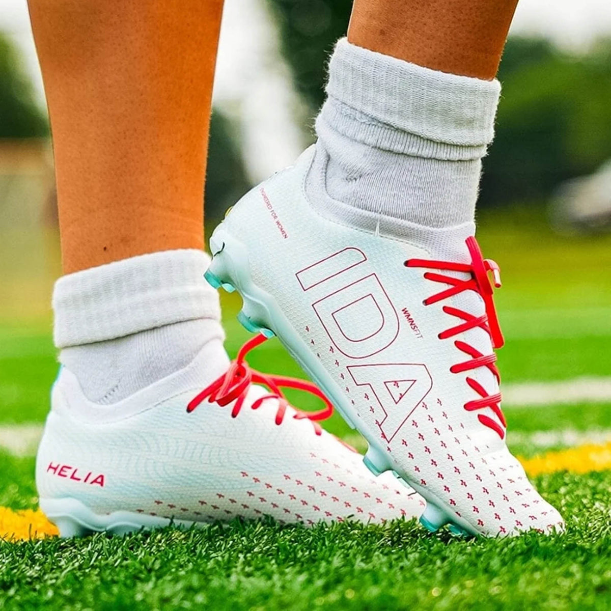 Close-up of a person wearing IDA Sports IDA Helia Club FG/AG soccer cleats in white and blue, standing on a green turf field.