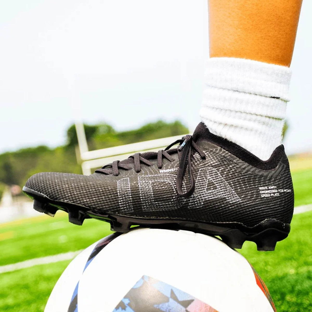 A person wearing IDA Sports IDA Helia Elite FG/AG cleats in Black/Dark Grey and white socks stands with one foot resting on a soccer ball on a grassy field.