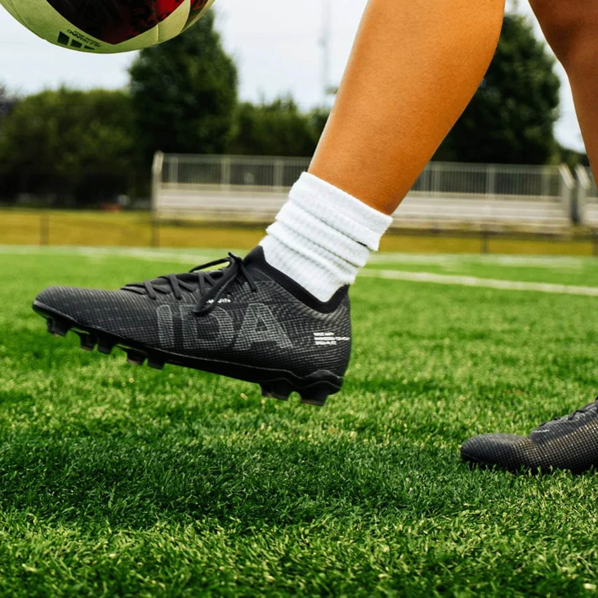 Close-up of a person wearing IDA Sports IDA Helia Elite FG/AG cleats in Black/Dark Grey with white socks, poised to kick a soccer ball on vibrant green artificial turf.