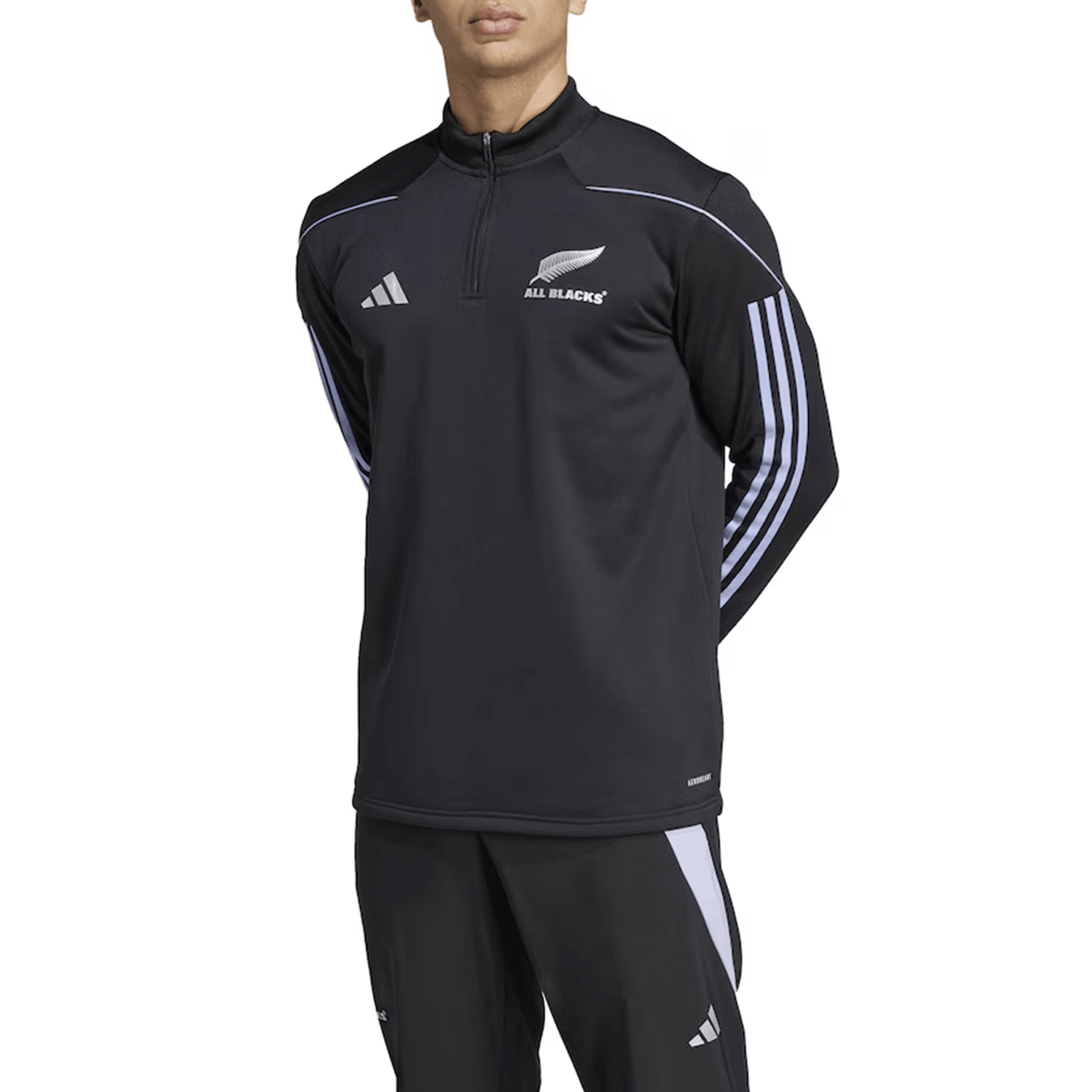 A person wearing the adidas All Blacks 24/25 Fleece 1/4 Zip and matching pants stands facing forward, hands behind their back, against a white background.