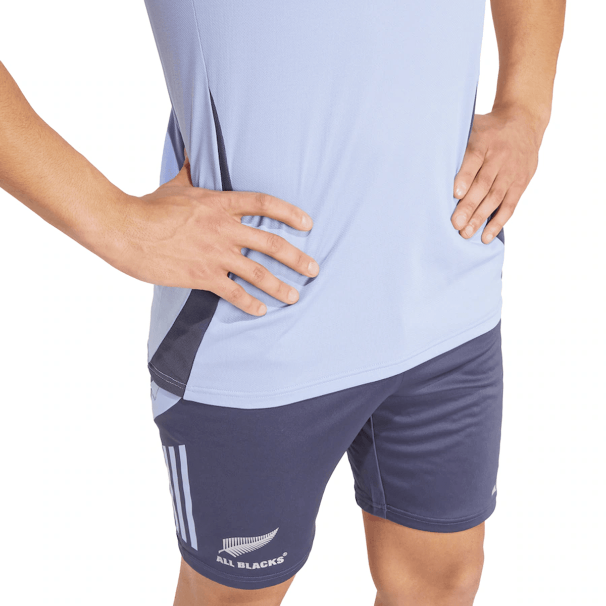 A person wearing the All Blacks 24/25 Performance Polo by adidas and navy shorts, proudly displaying the "All Blacks" logo, stands confidently with hands on hips, embodying the spirit of the New Zealand rugby team.