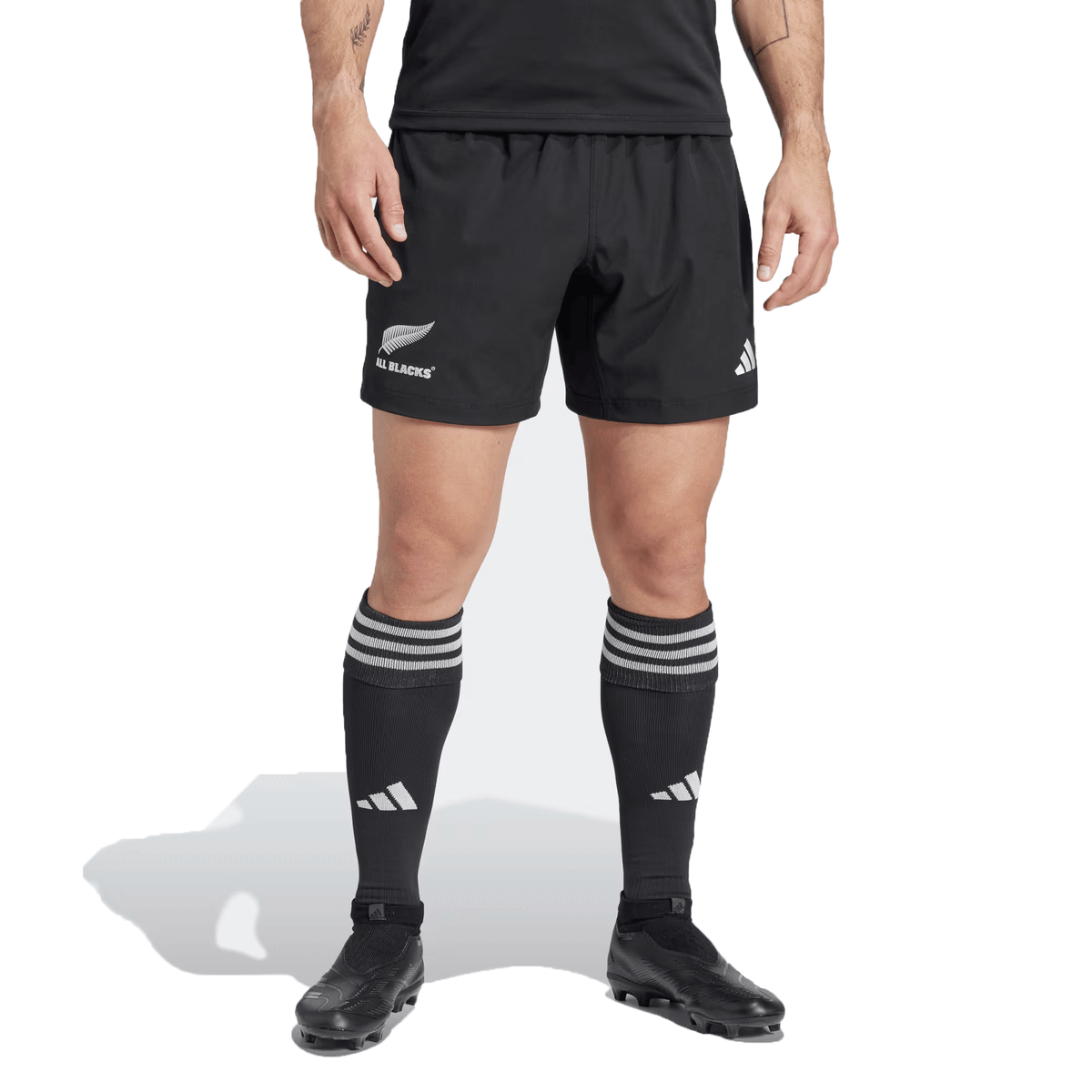 A person wearing the All Blacks 24/25 Home Shorts by adidas, crafted from recycled materials, with matching socks and cleats, stands against a plain background.