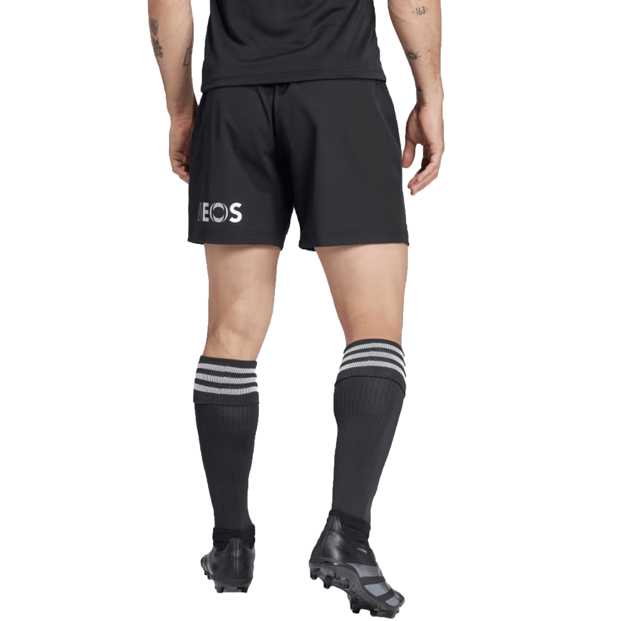 A person in a black sports uniform, wearing the All Blacks 24/25 Home Shorts by adidas made from recycled materials, paired with black socks featuring white stripes and black cleats, stands on a white background.