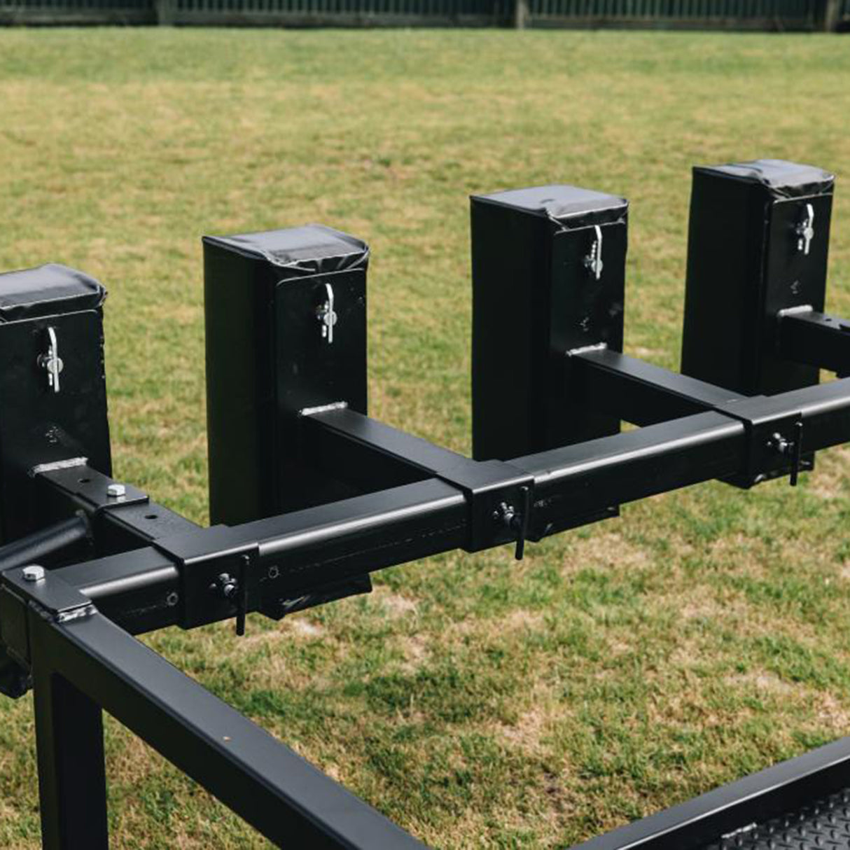 A row of four black metal lockboxes, part of the Iron Back Gorilla 8 Man Scrum Machine by IronBack, is mounted on a sturdy metal frame and stands on a grassy lawn, evoking the robust design of scrum training equipment, with a wooden fence visible in the background.