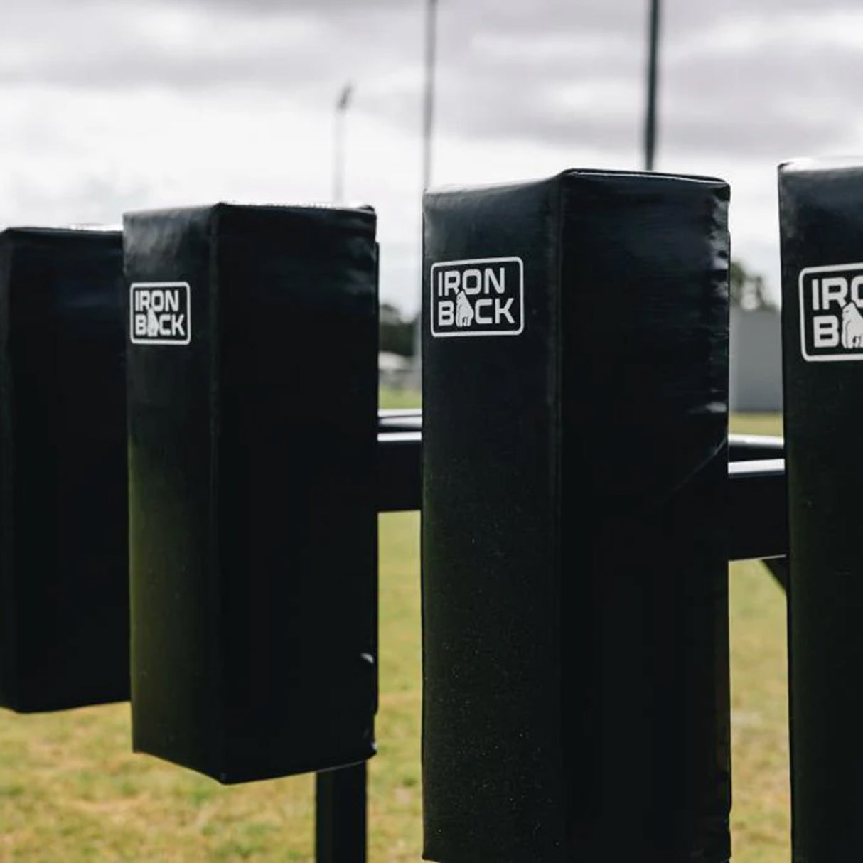 Four black padded blocking dummies, part of the Iron Back Gorilla 8 Man Scrum Machine by IronBack, are arranged outdoors on a grassy field under a cloudy sky as essential scrum training equipment.