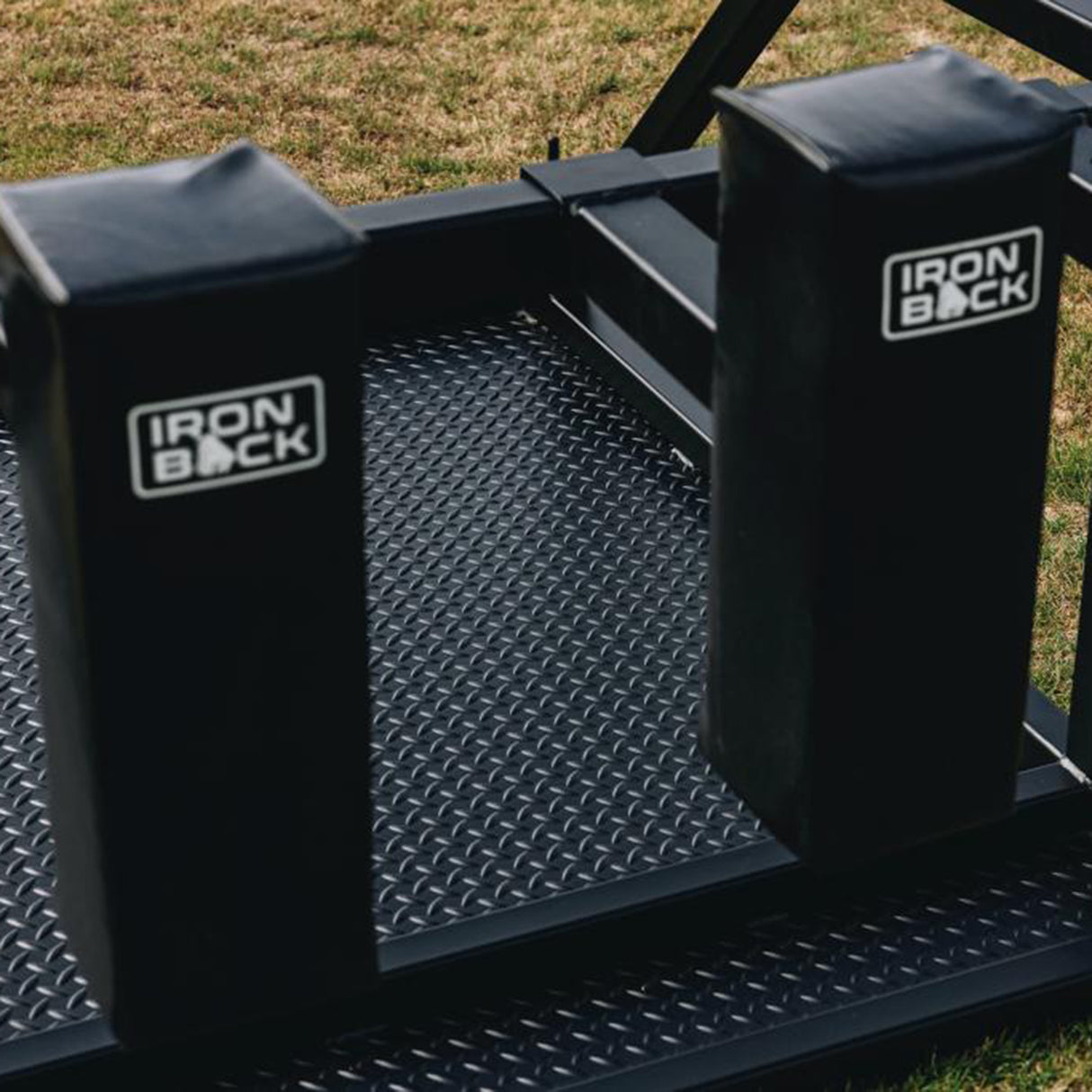 The IronBack Gorilla 8 Man Scrum Machine features a rugged black metal frame with a diamond plate surface and two padded blocks branded "IronBack." Designed for durability and effective team practice, this scrum training equipment is perfect for rugby drills and is shown placed outdoors on grass.