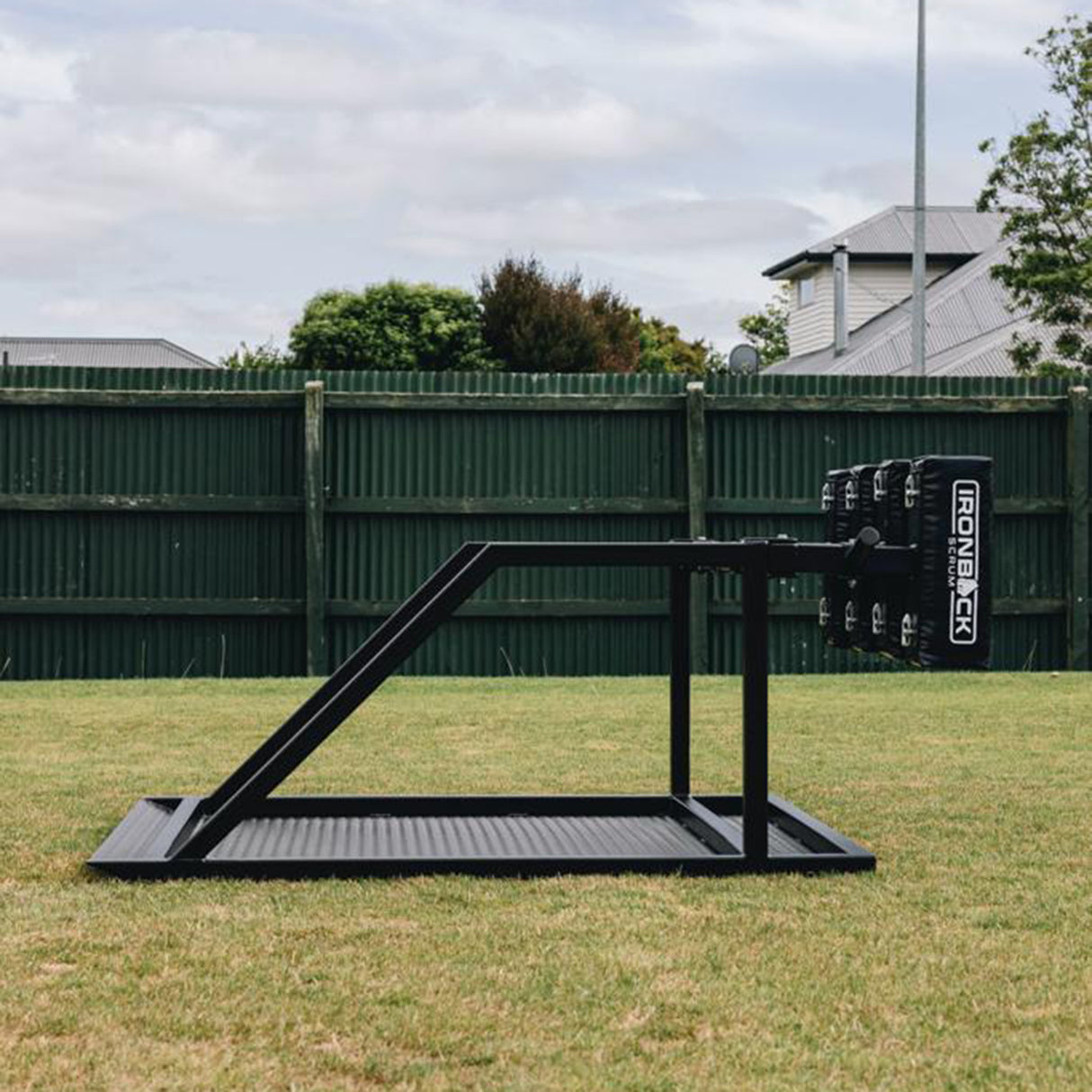 The IronBack Iron Back Gorilla 8 Man Scrum Machine—black metal scrum machine positioned on grass in a fenced backyard, with houses and trees in the background—is ideal for building strength and technique during rugby scrum training.
