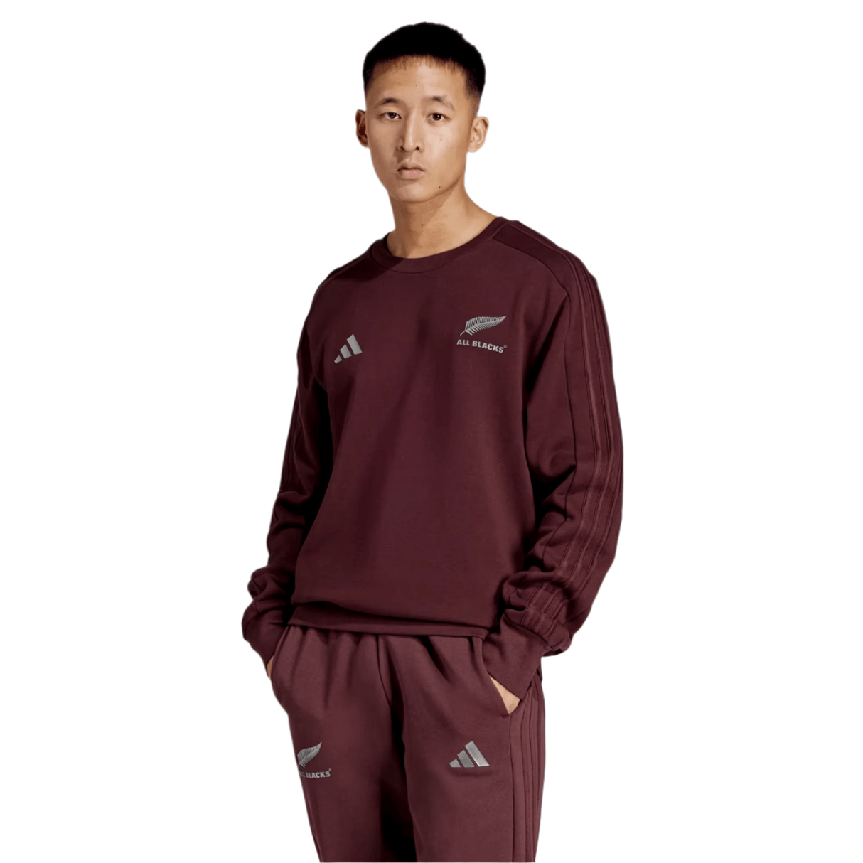 A person in an All Blacks 24/25 Fan Crewneck Sweatshirt by adidas and logo-adorned pants stands confidently against a plain background.