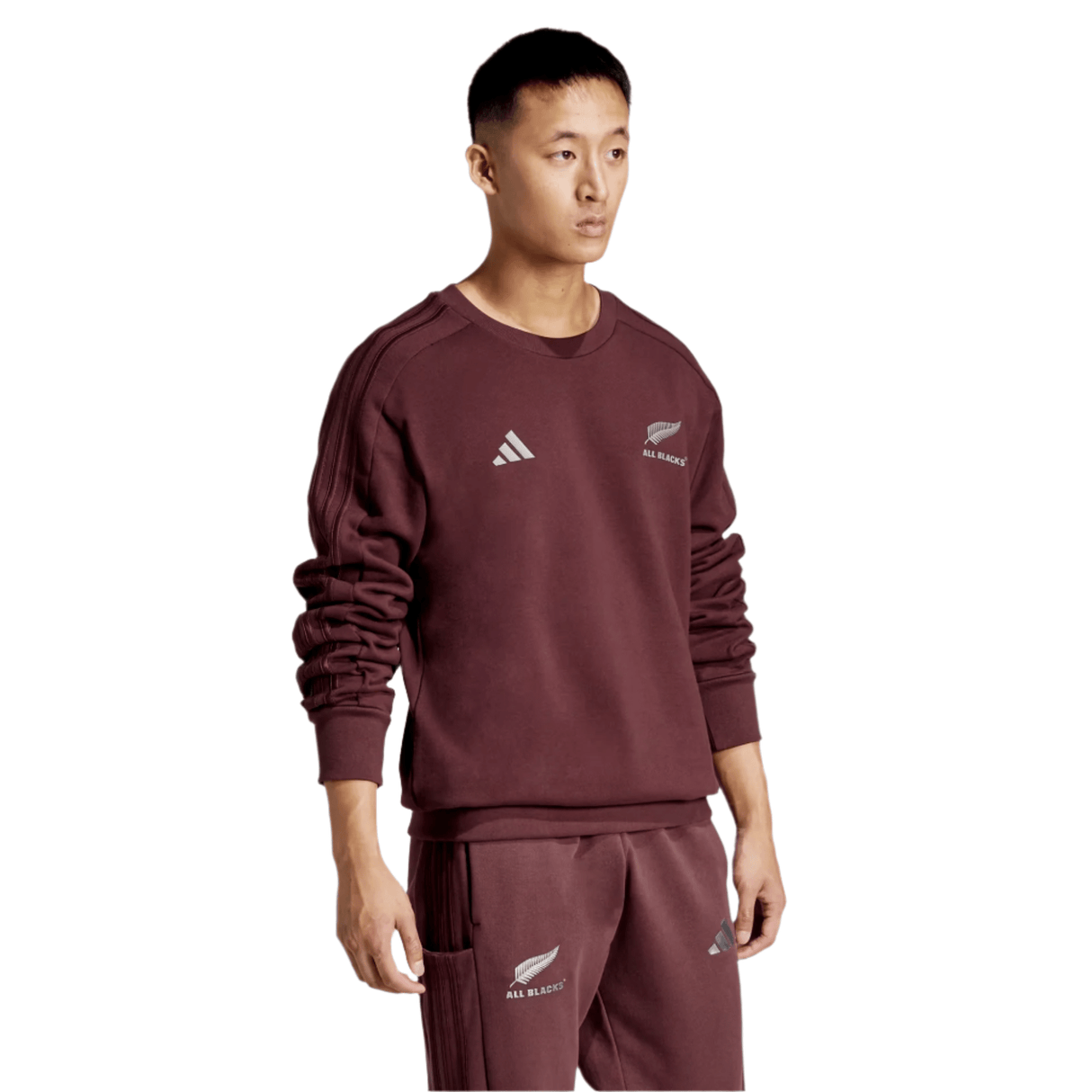 Clad in an All Blacks 24/25 Fan Crewneck Sweatshirt by adidas and matching pants, a person stands poised against a plain backdrop, both items emblazoned with the iconic All Blacks and adidas logos.
