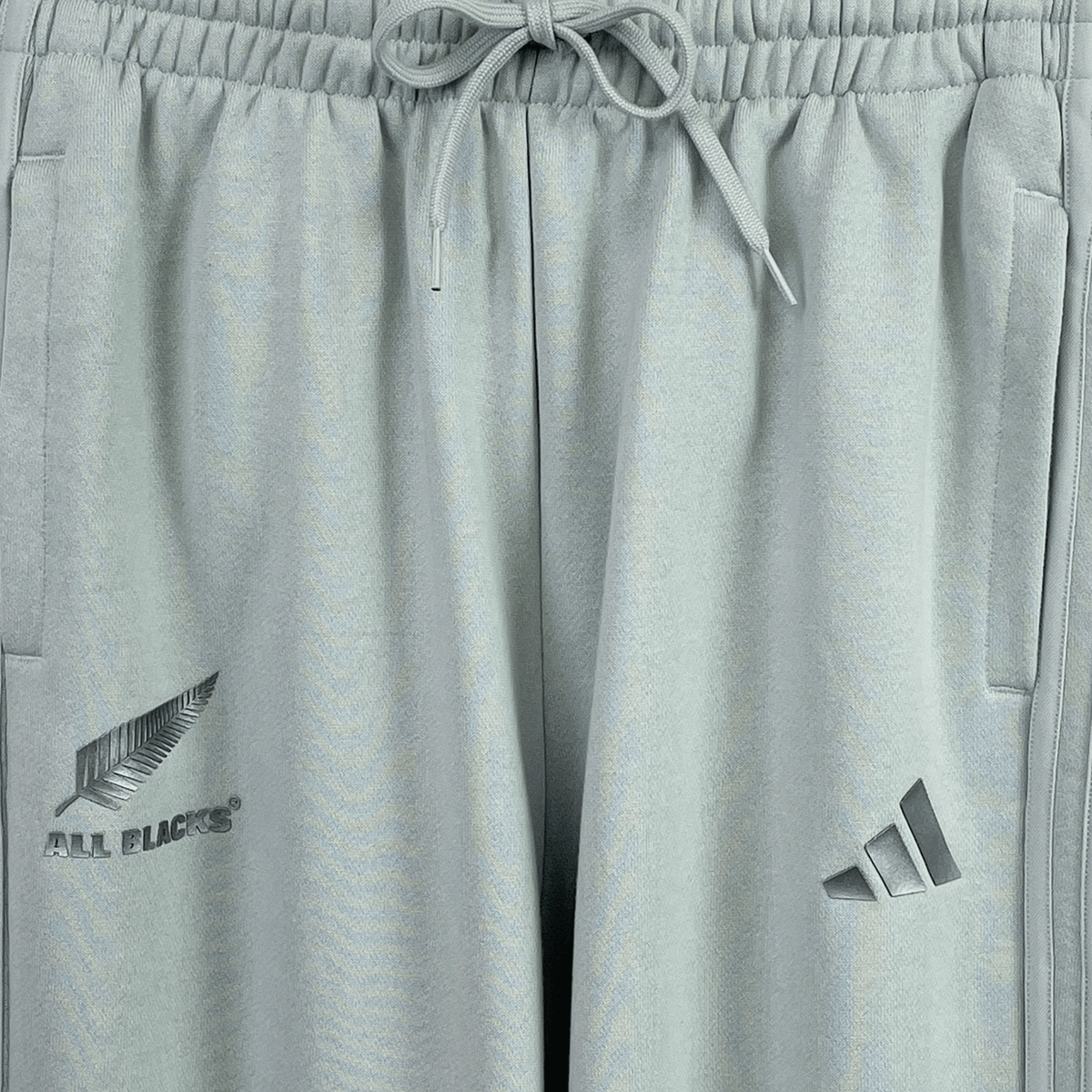 The Women's All Blacks 24/25 Fan Cuffed Pant by adidas features gray sweatpants with a drawstring waist, adorned with the silver fern and "All Blacks" logo on the left and the adidas fan wear logo on the right, ideal for New Zealand rugby fans.