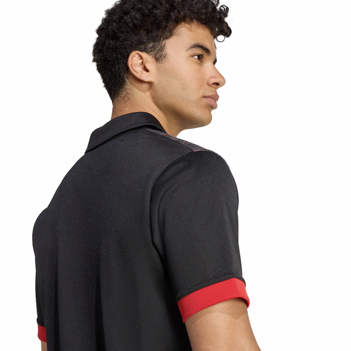 A young man with curly hair is seen from the back and side, wearing the adidas Maori All Blacks 25/26 Replica Home Jersey with red sleeve trim, against a plain white background.
