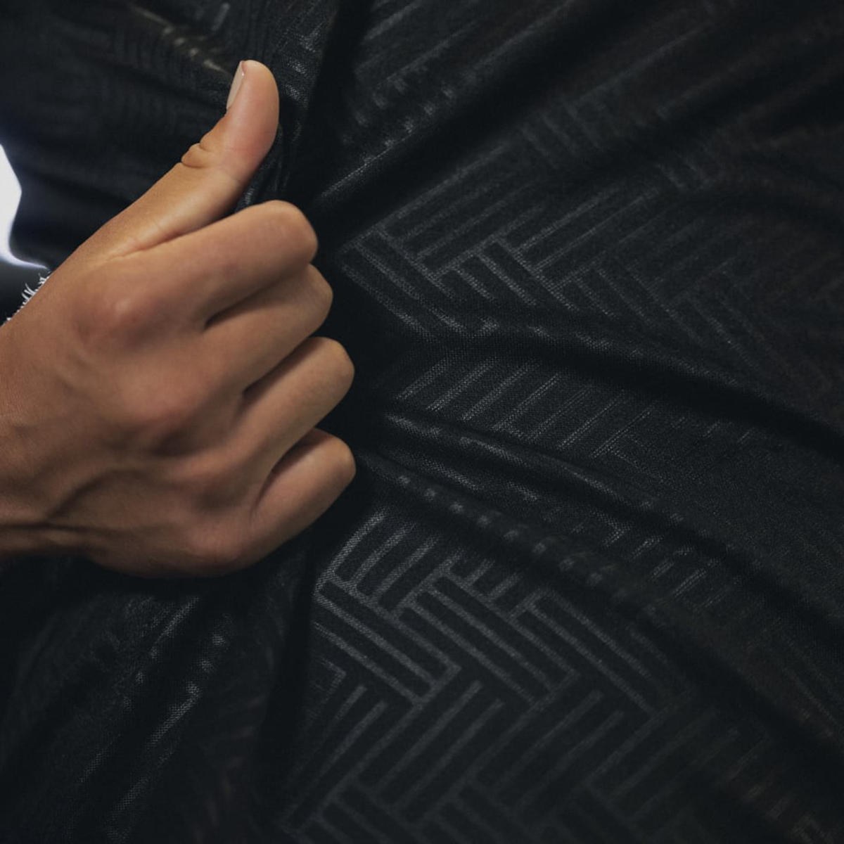 A hand grips a dark fabric with a subtle geometric pattern, evoking the adidas Women's Black Ferns WRWC25 Replica Home Jersey.