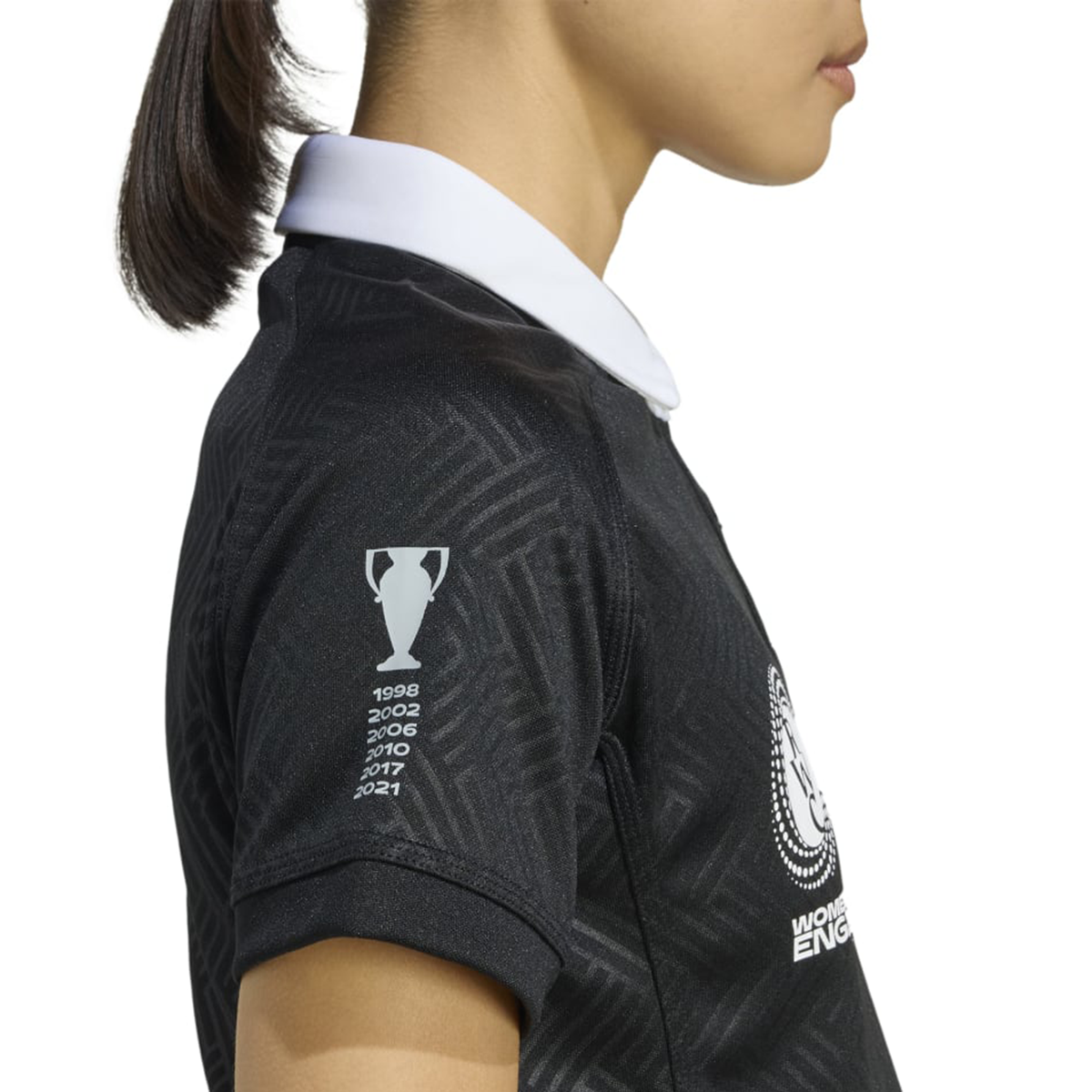 A person wears the Women's Black Ferns WRWC25 Replica Home Jersey by adidas, a black collared sports shirt with a white collar and sleeve trophy graphic listing years, inspired by the official Black Ferns kit.