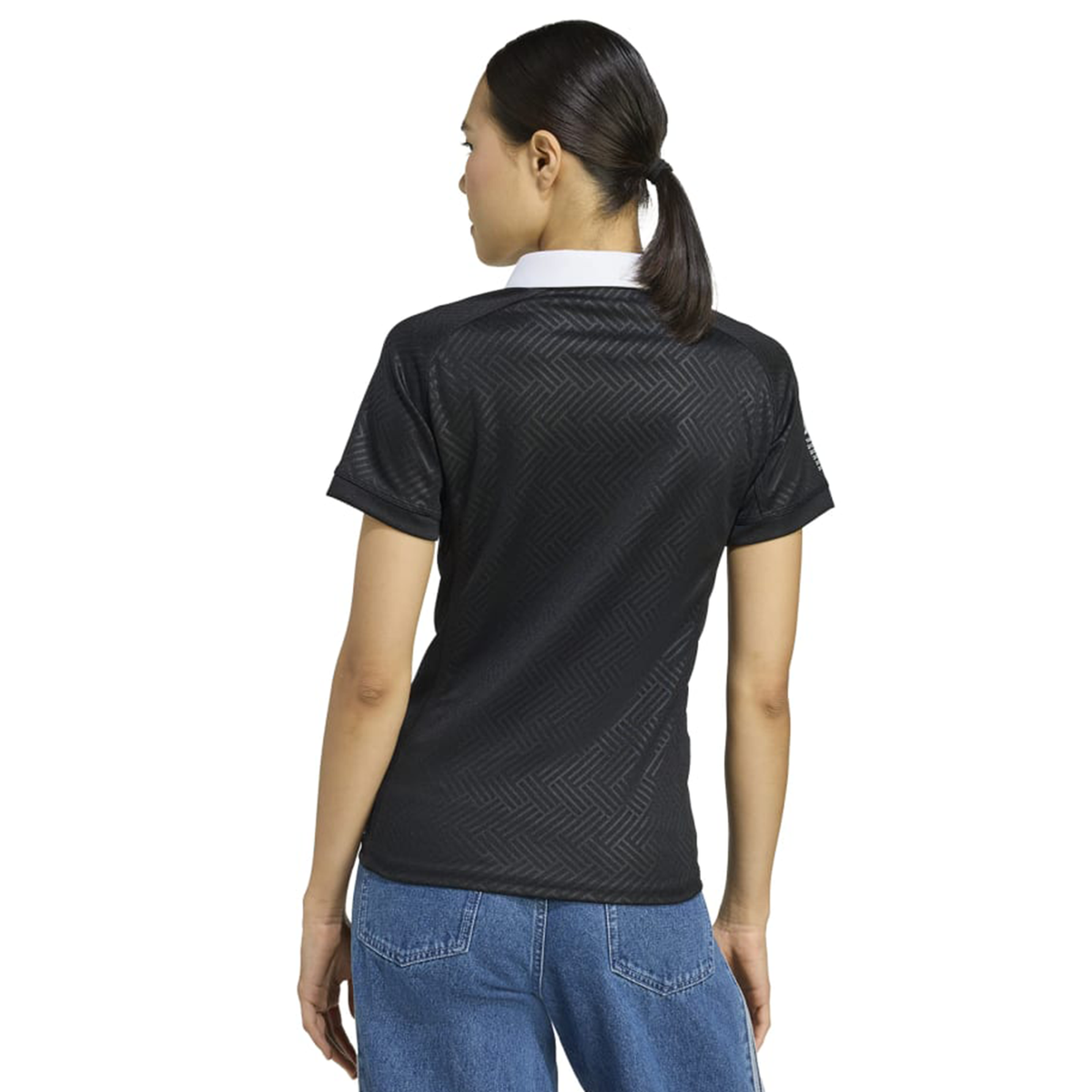 A woman with dark hair tied back is seen from the back wearing the Women's Black Ferns WRWC25 Replica Home Jersey by adidas and blue jeans.