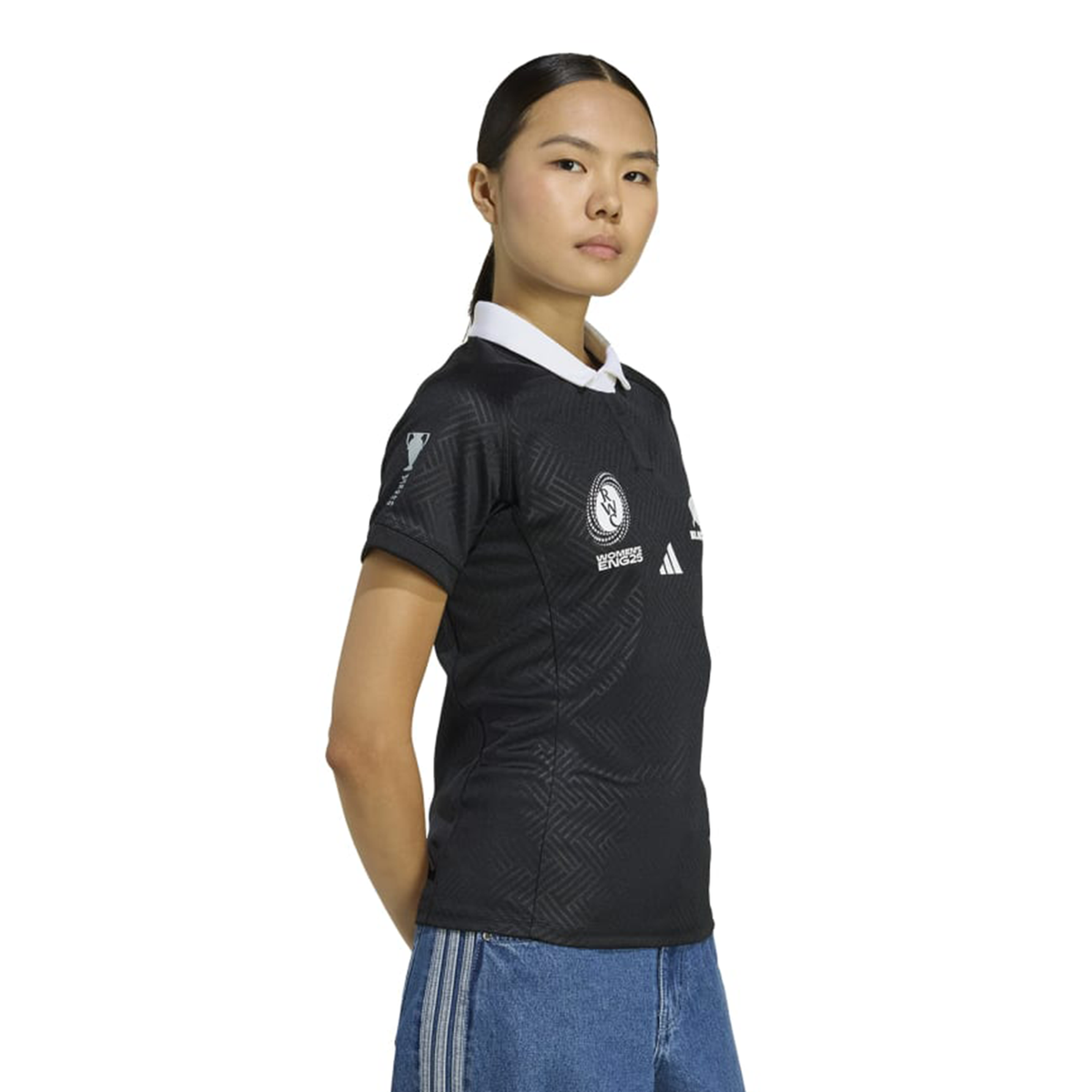 A woman stands with hands behind her back, wearing the adidas Women's Black Ferns WRWC25 Replica Home Jersey and blue jeans against a plain background.