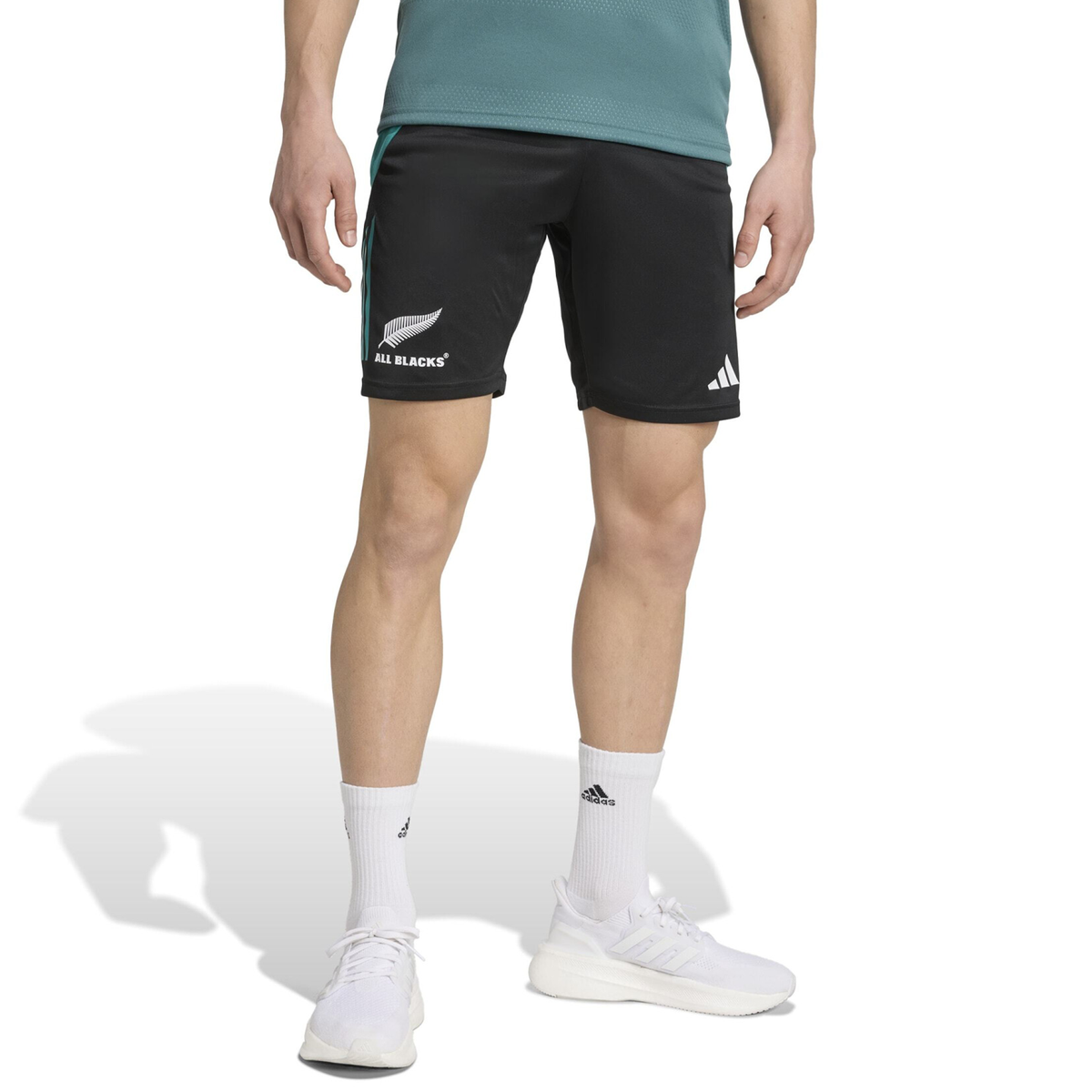 A person wears the adidas All Blacks 25/26 Training Gym Short, a teal shirt, white adidas socks, and white adidas sneakers against a white background, displaying their New Zealand rugby style.